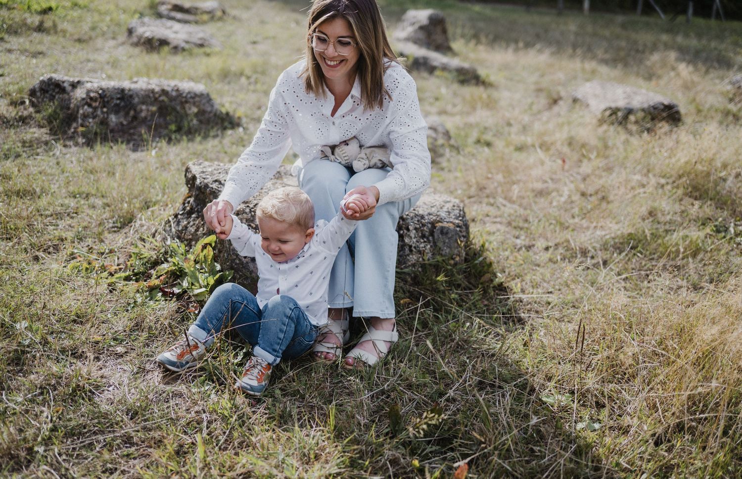 A smiling woman sits on a rock outdoors with a toddler in a grassy field on a sunny day.