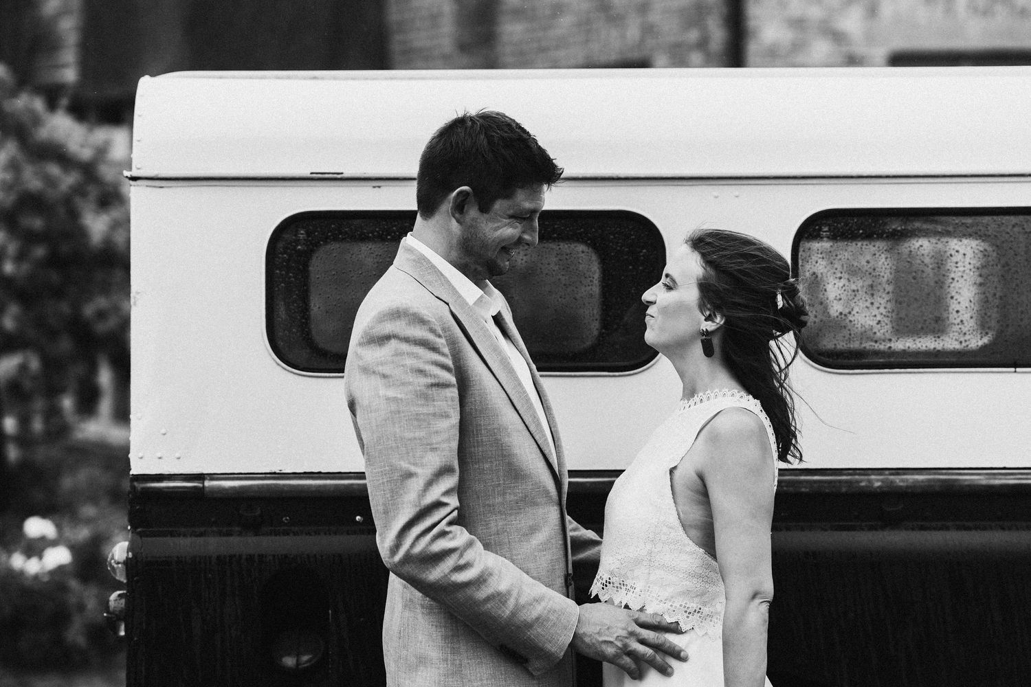 Black and white photo of a smiling couple in wedding attire standing in front of a vintage vehicle.