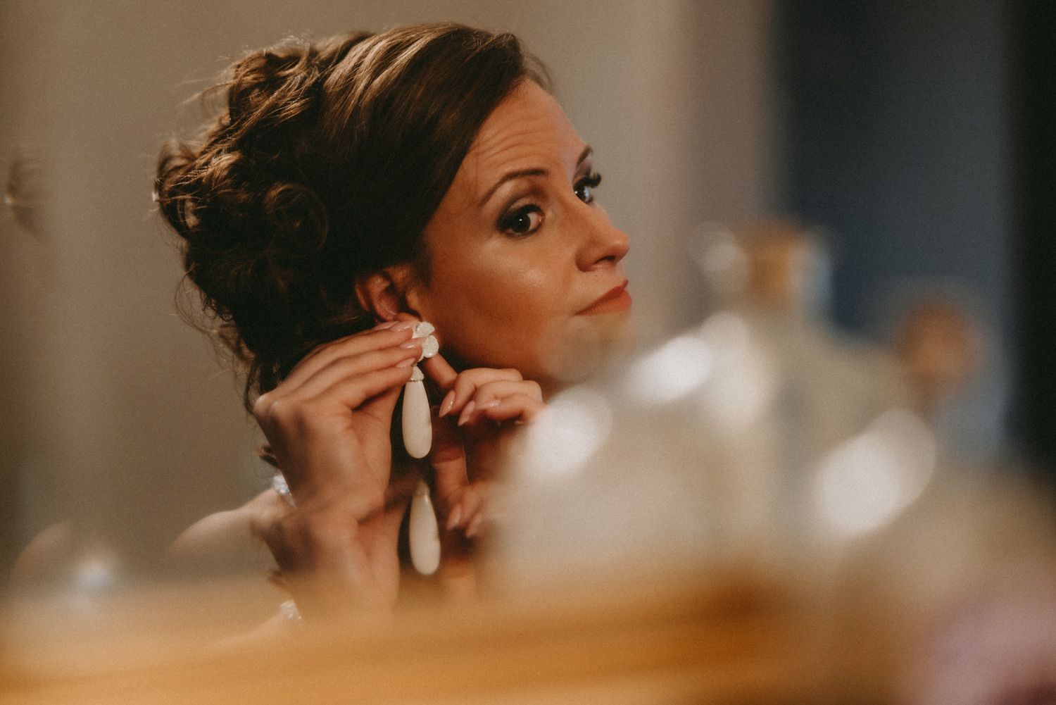 Bride putting on elegant drop earrings while getting ready, reflected in a mirror on her wedding day.