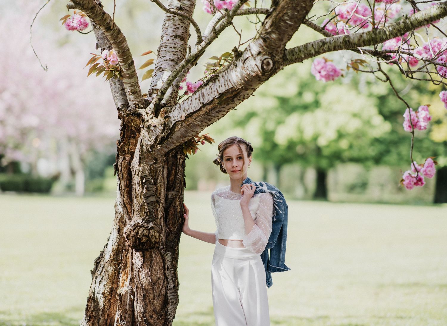 Bride in white outfit leaning against cherry blossom tree in lush green park, holding blue jacket.