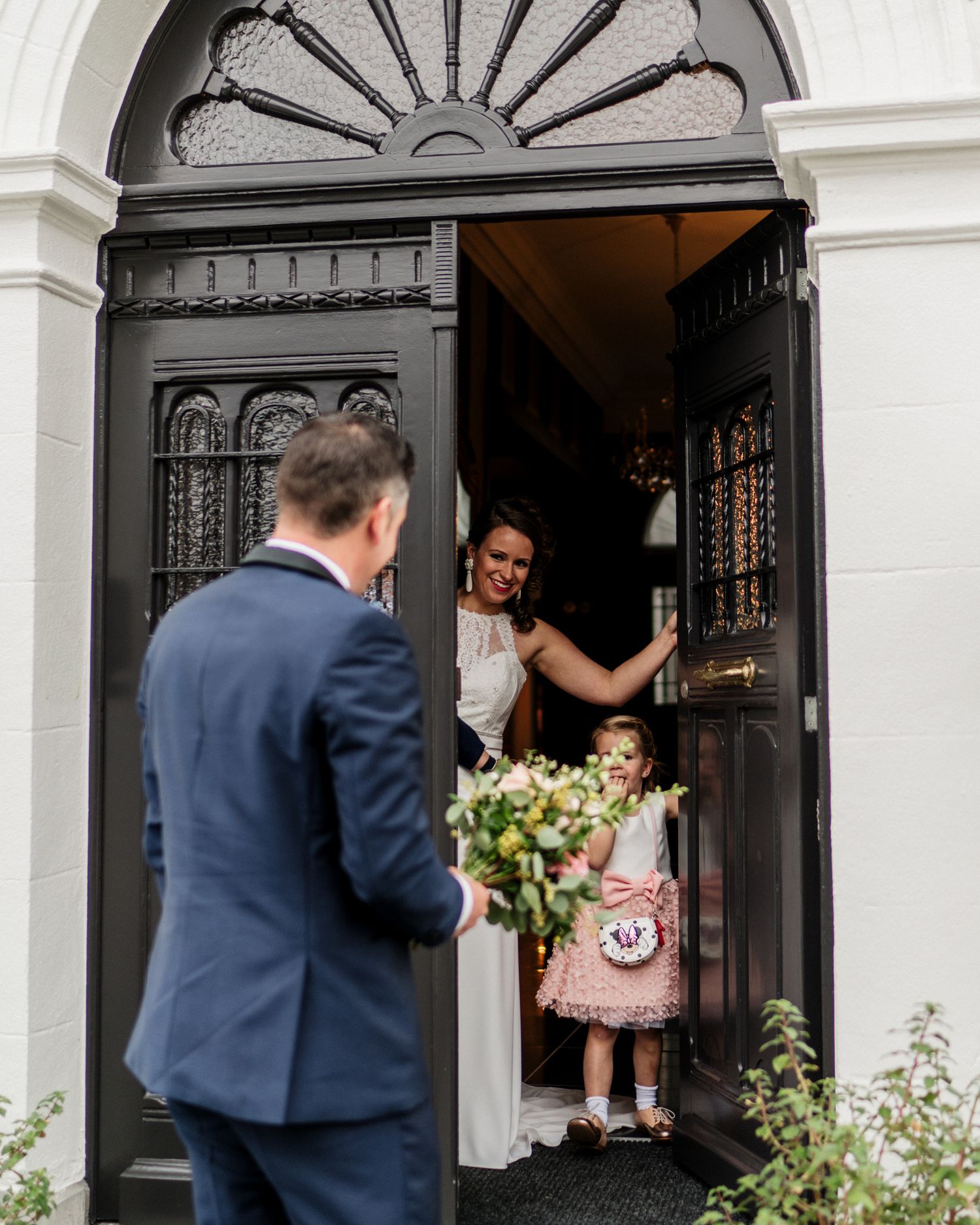 Bride and flower girl greet groom at elegant black door during wedding day first look ceremony.