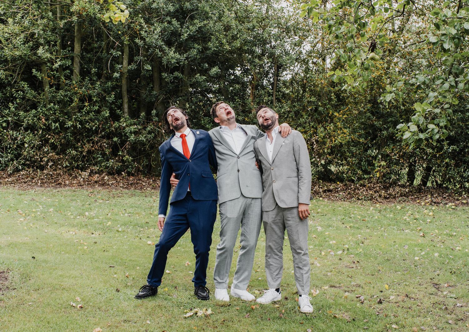 Three men in suits laughing and posing together outdoors on grass with green trees in the background.