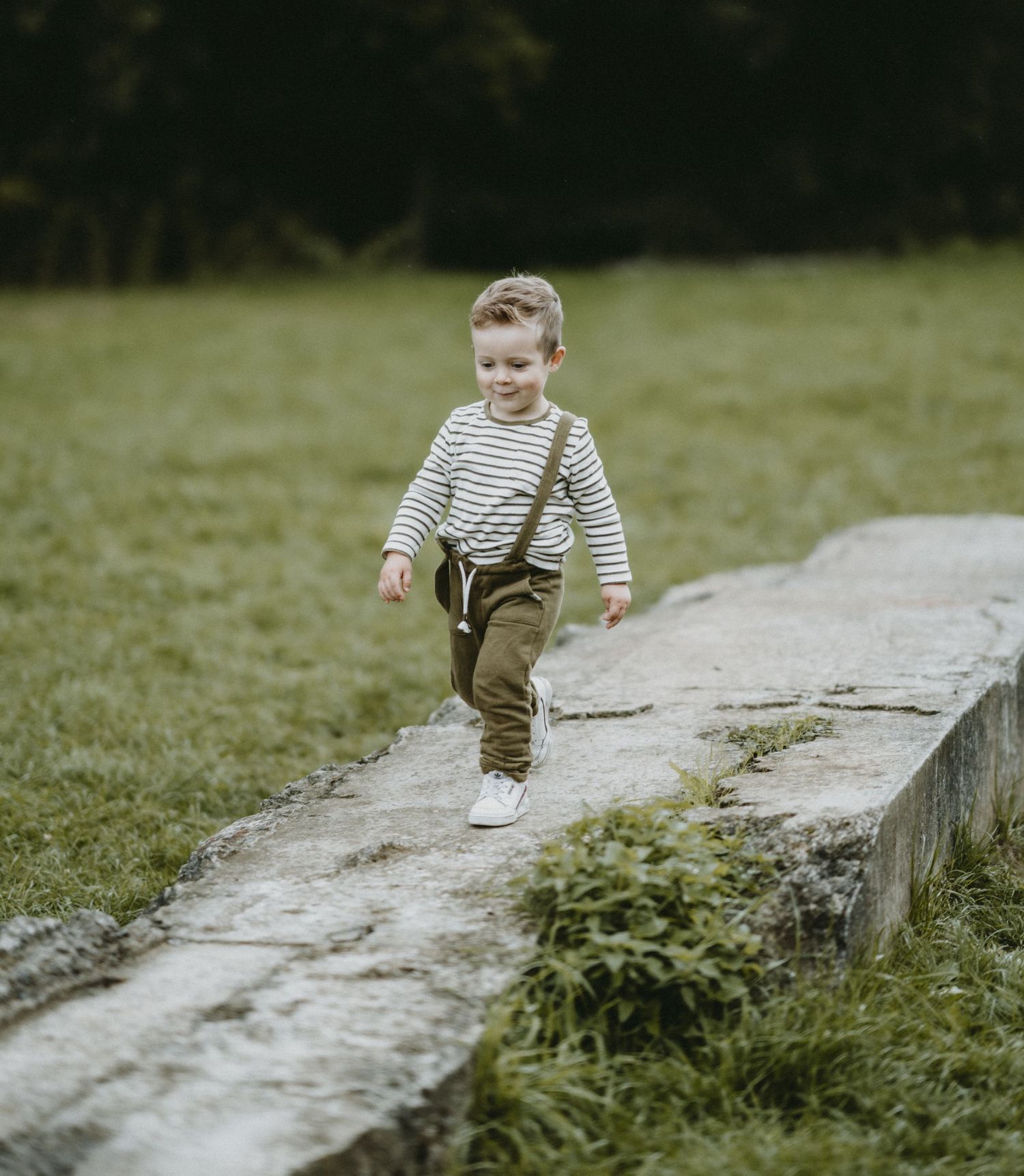 A child in striped shirt and brown pants runs along a concrete path in a grassy field.