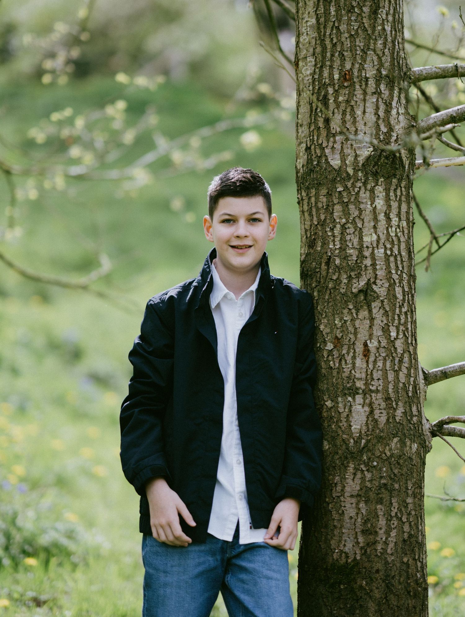 A teen in a navy jacket and white shirt poses casually against a tree trunk in a spring garden.