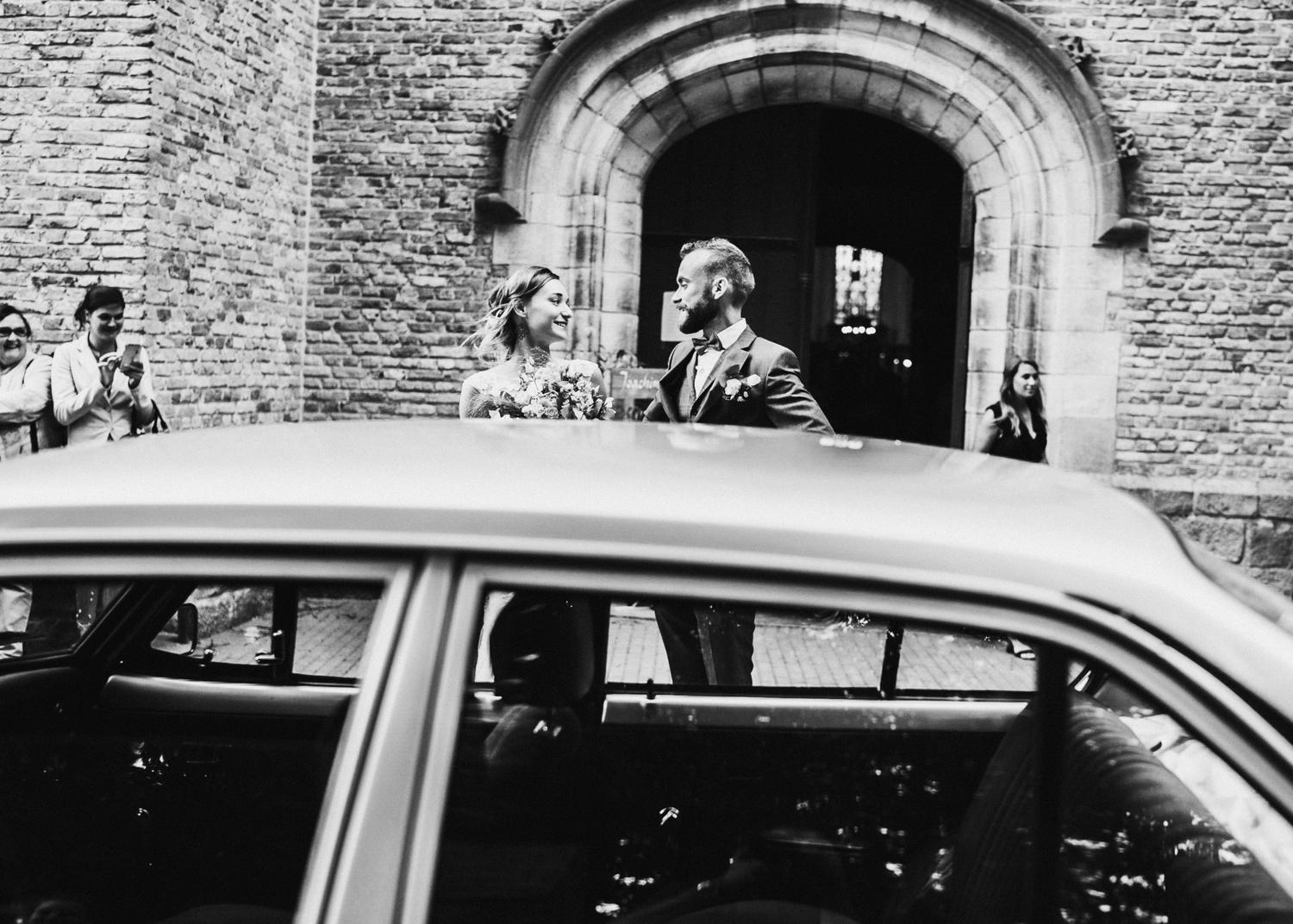 Black and white photo of a bride and groom leaving a stone church, smiling beside a vintage wedding car.