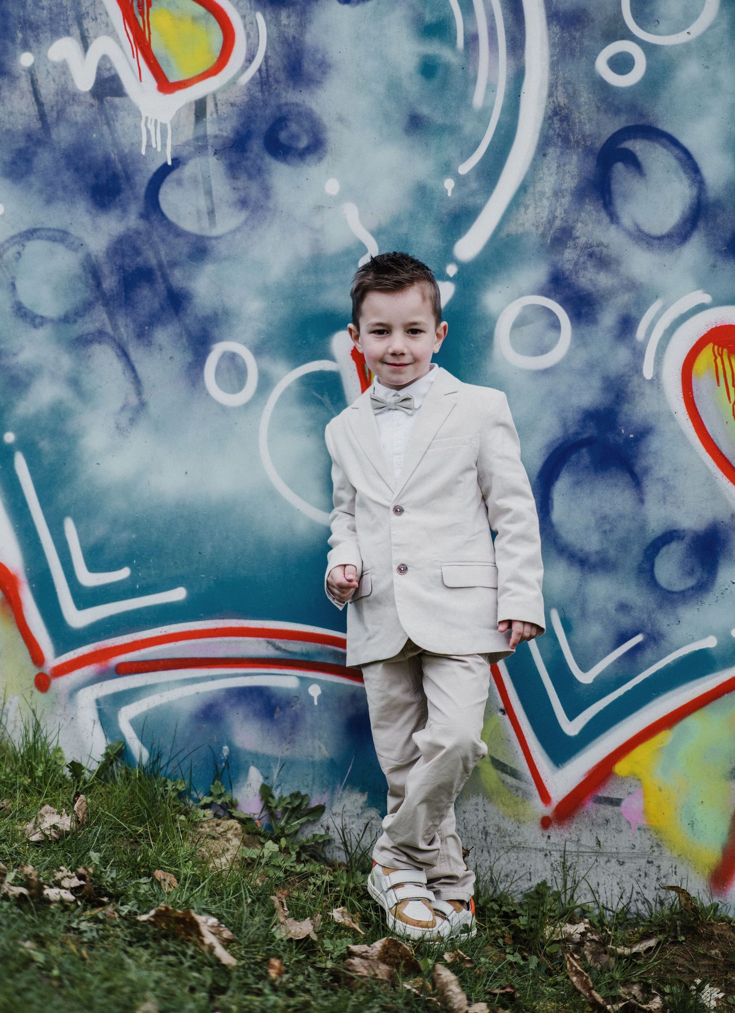 A young boy in a white suit and bow tie poses confidently in front of a colorful graffiti wall outdoors.