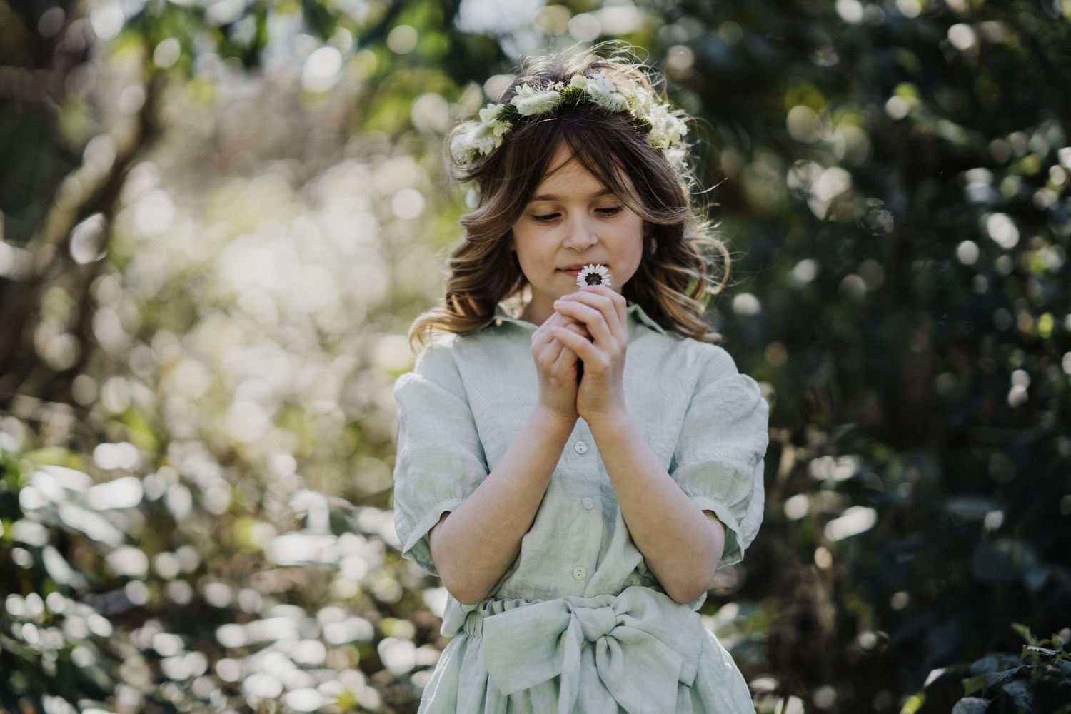 Young girl in floral crown smells white wildflower in sunlit garden, wearing sage green dress.