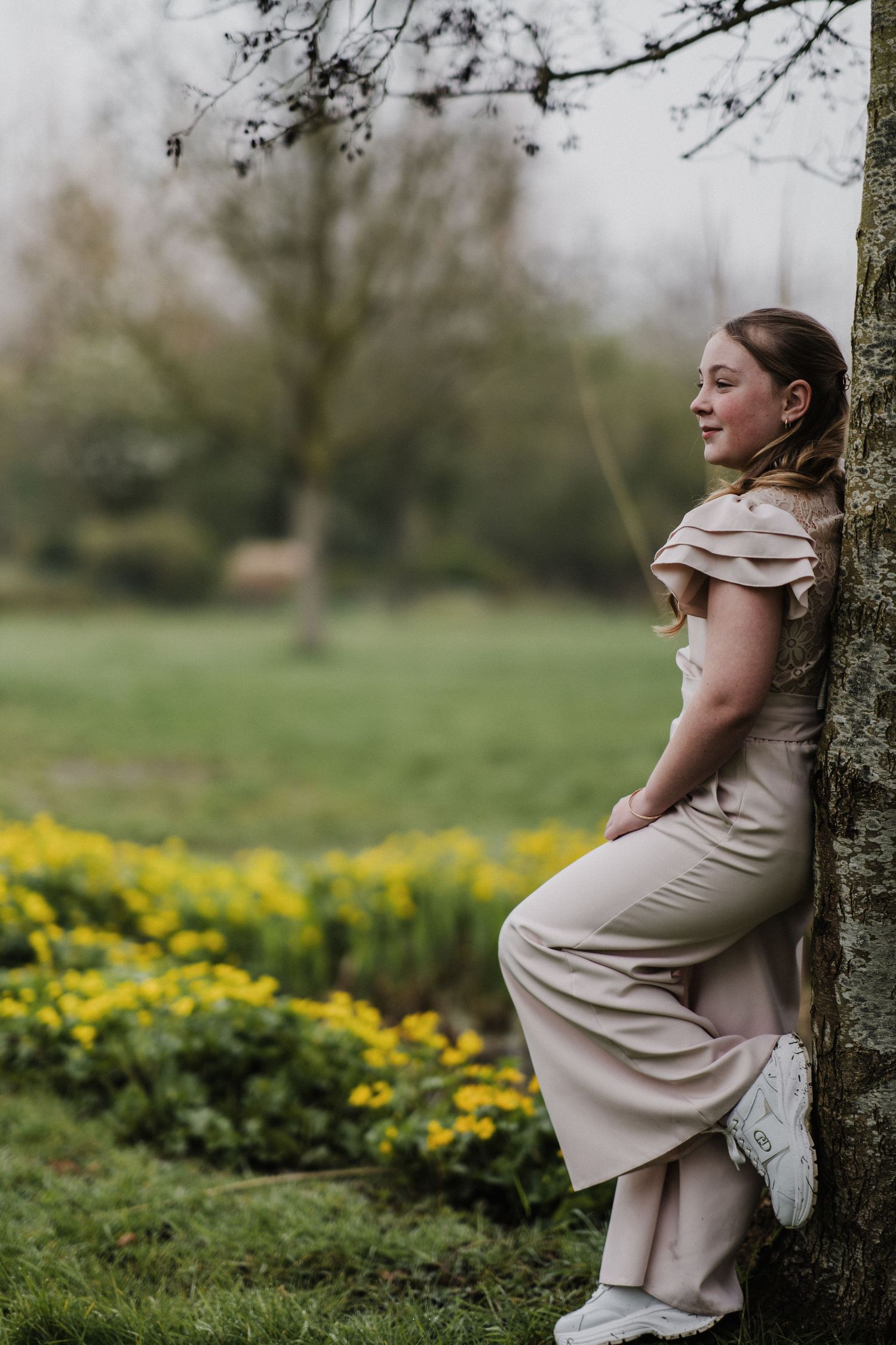 Young girl in pink dress leaning against tree in lush green park with yellow flowers blooming in background.