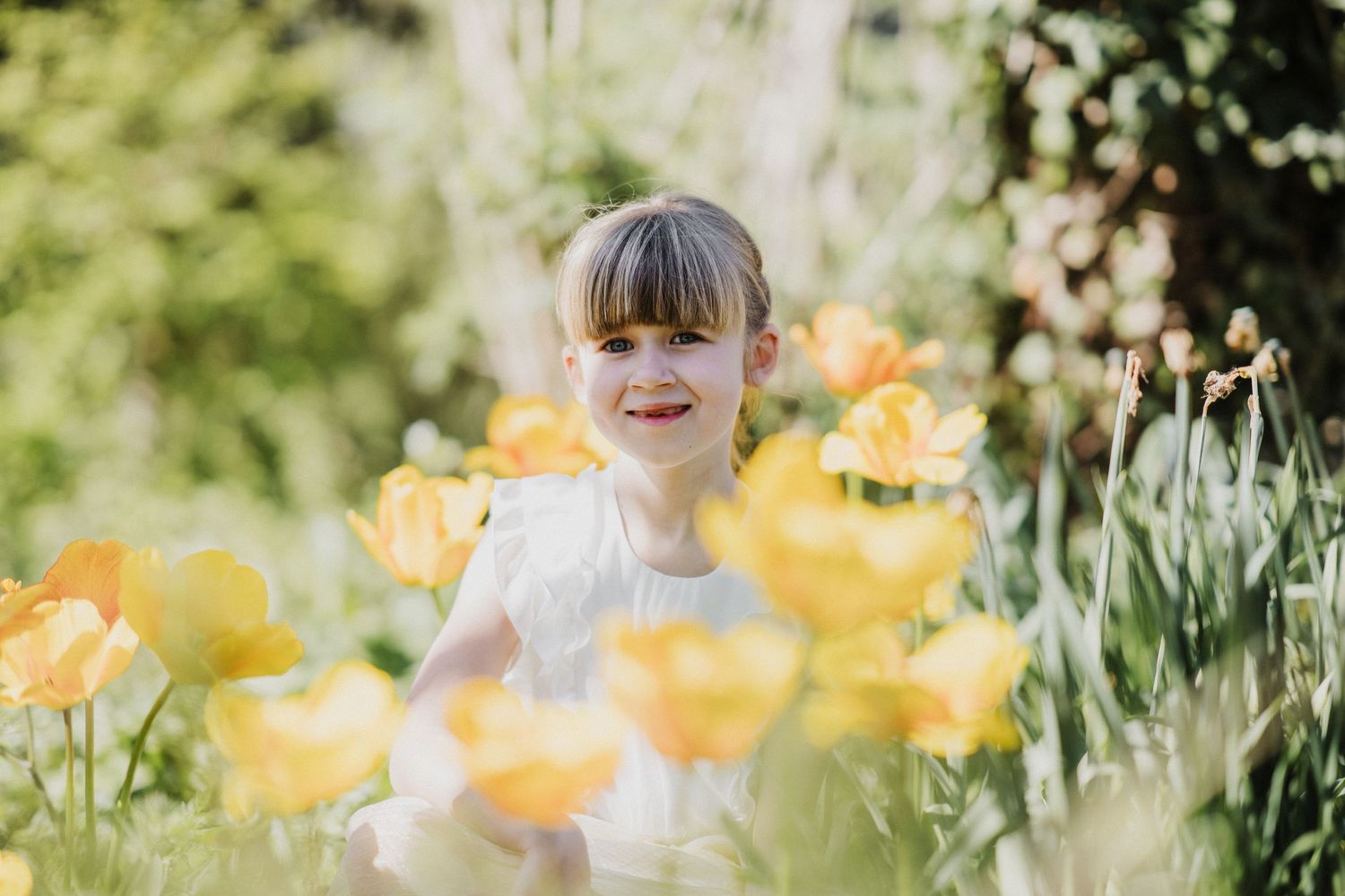 A young girl in a white dress smiles among vibrant yellow tulips in a sunny garden.