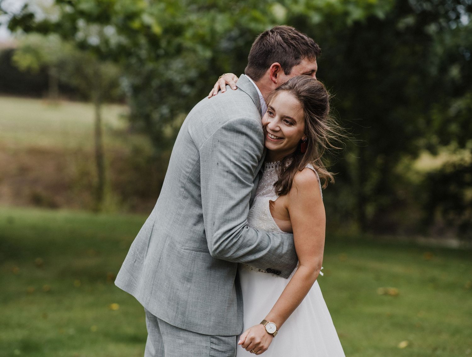 A bride and groom share a romantic embrace outdoors, smiling joyfully in wedding attire on a lush green lawn.