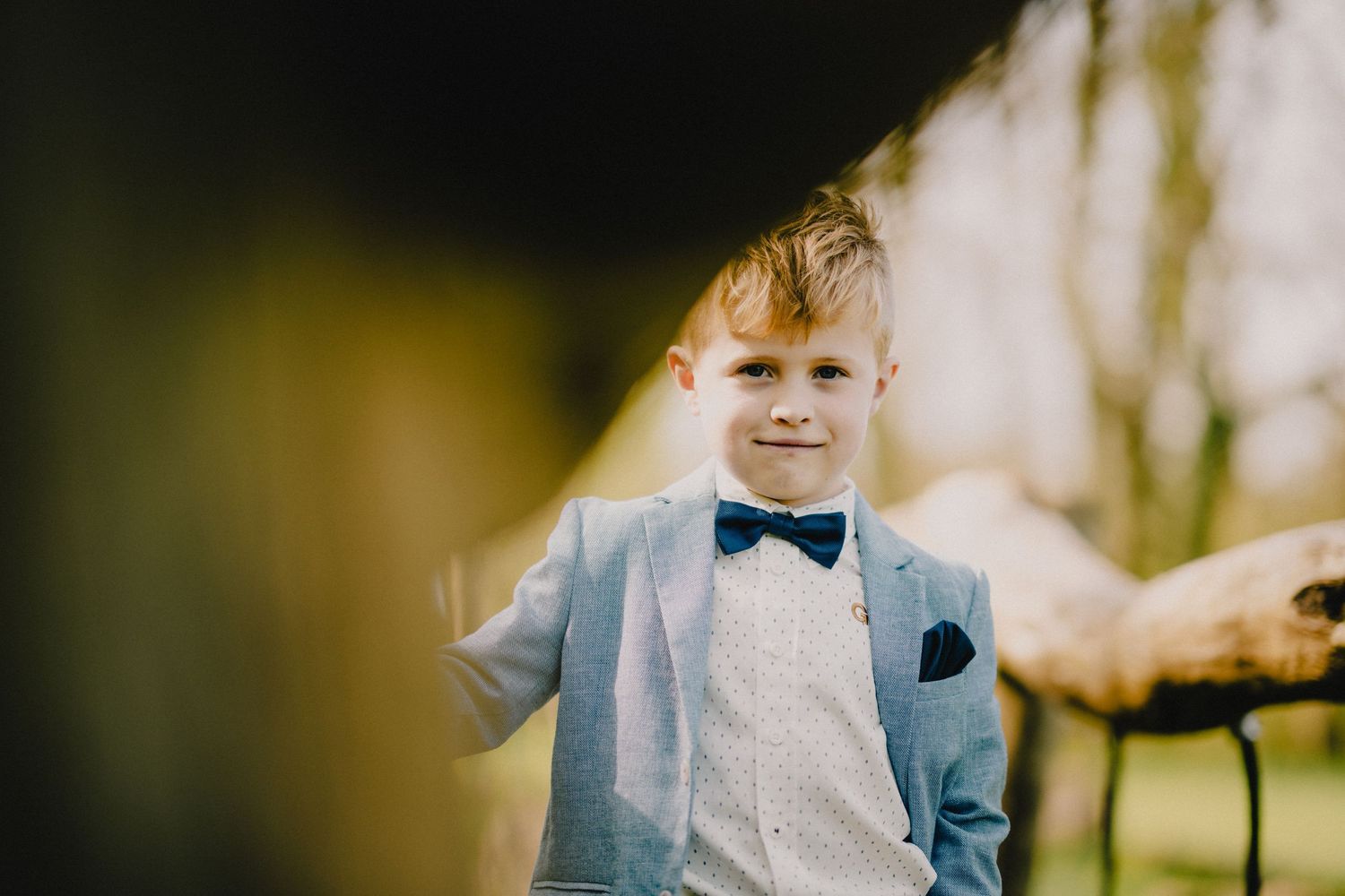 A young child in a light blue suit and bow tie smiles at the camera in an outdoor setting.