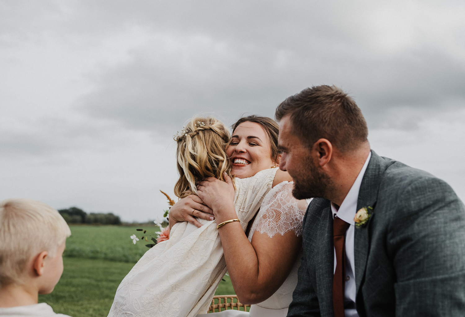 Bride and groom laughing and embracing a guest during outdoor wedding ceremony on a cloudy day.