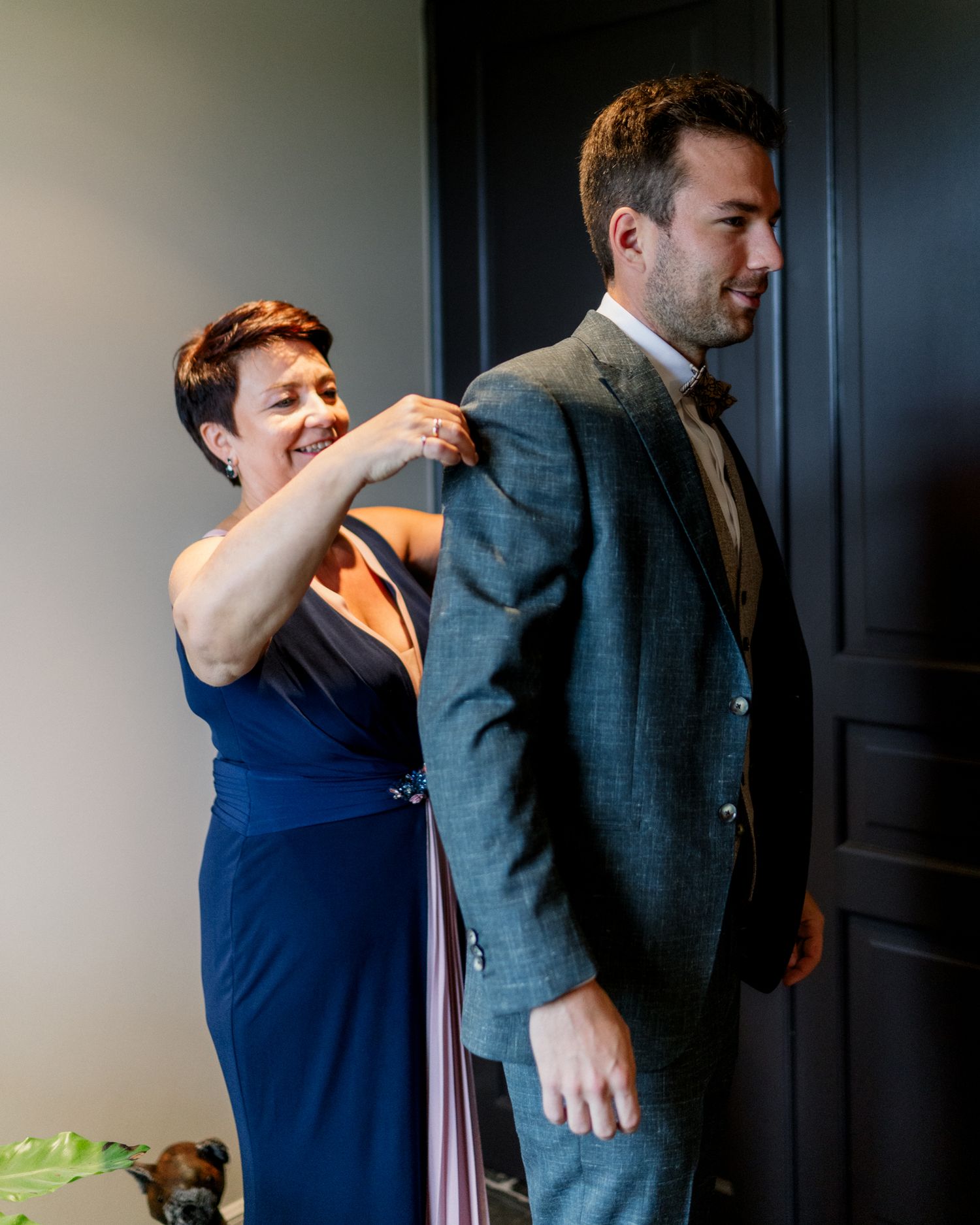 A woman in a blue dress adjusts the back of a man's gray suit jacket at a wedding.