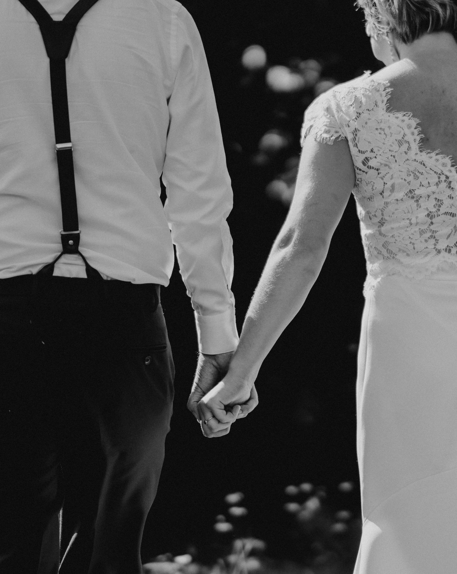 Black and white photo of a bride and groom holding hands, showcasing lace wedding dress and suspenders.