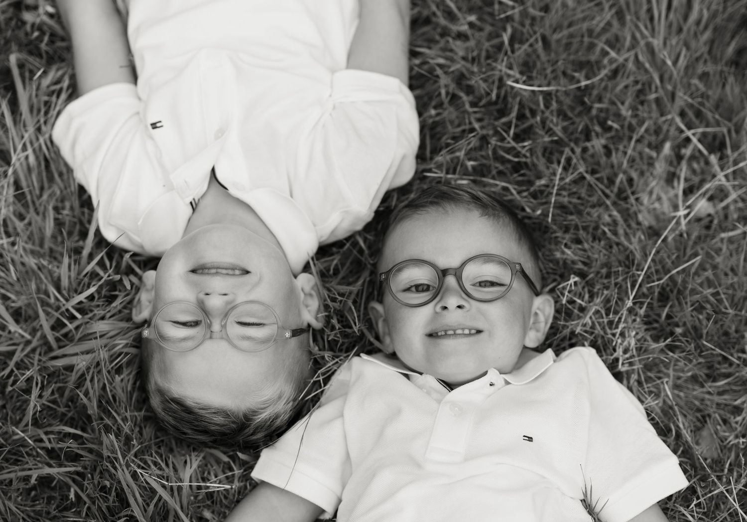 Two children wearing glasses lying in grass smiling, black and white photo.