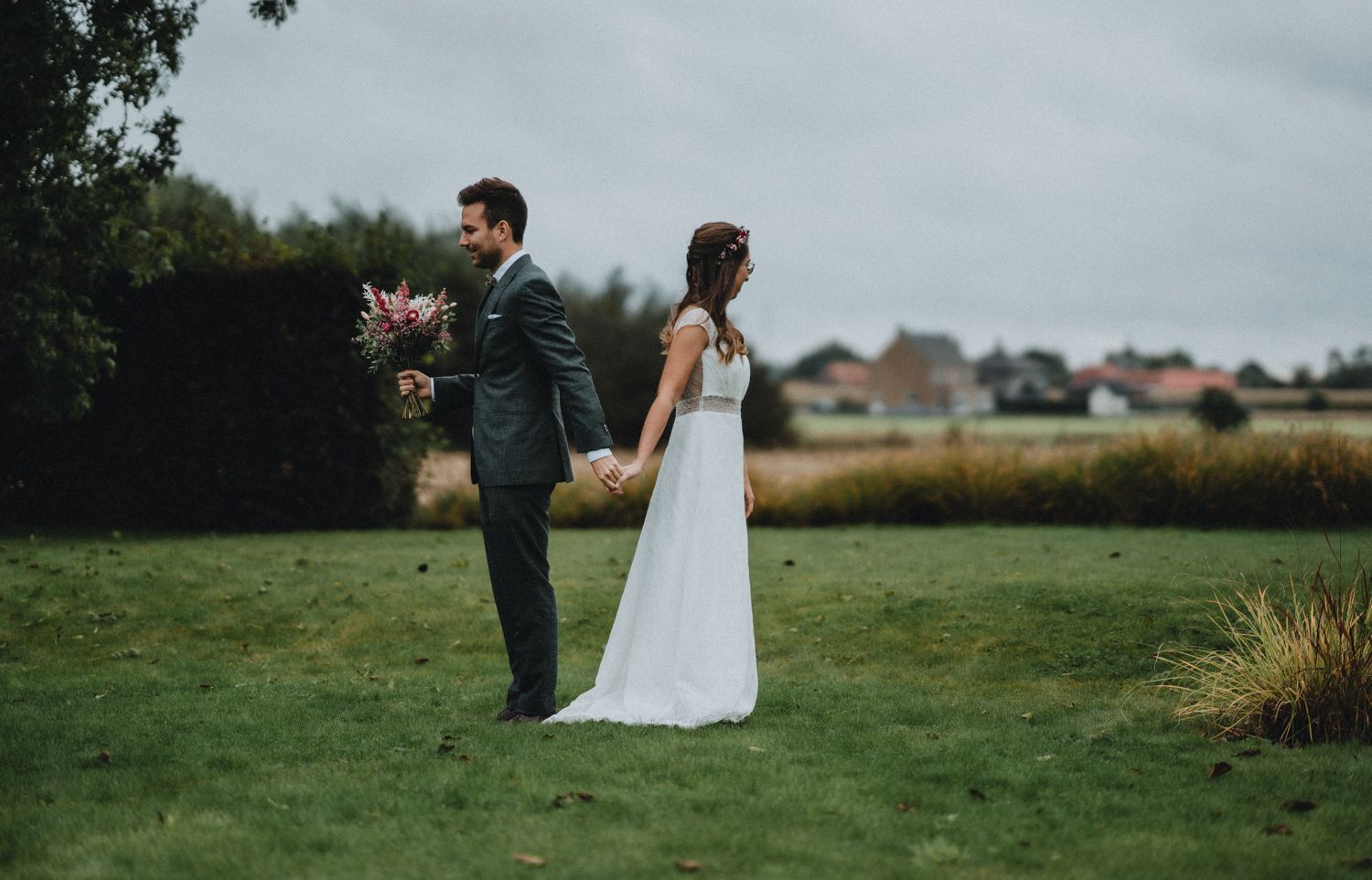 Artistic wedding photo of a couple standing back-to-back holding bouquets in a misty field.