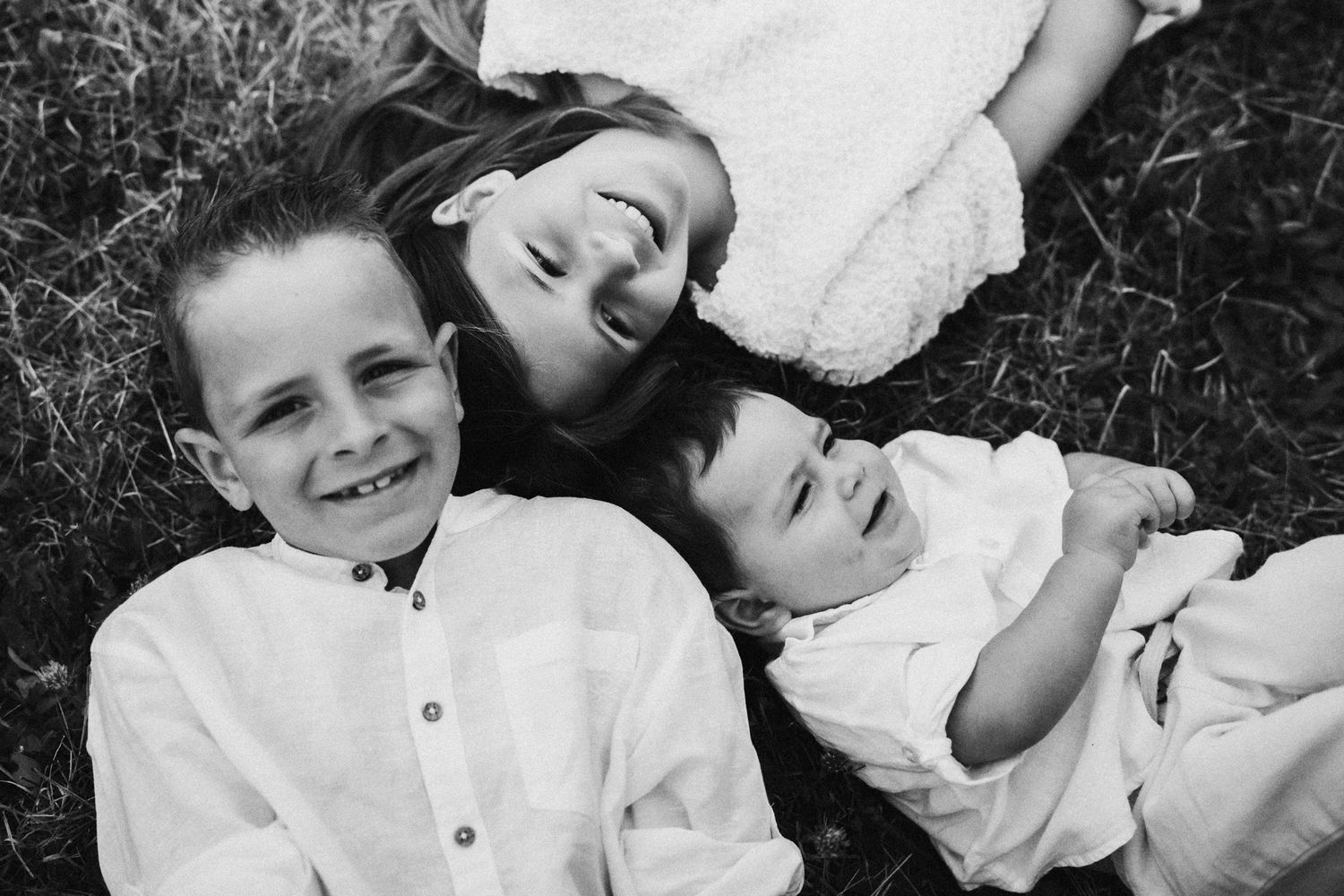 Black and white photo of three smiling children lying on grass, wearing white outfits in a candid family portrait.