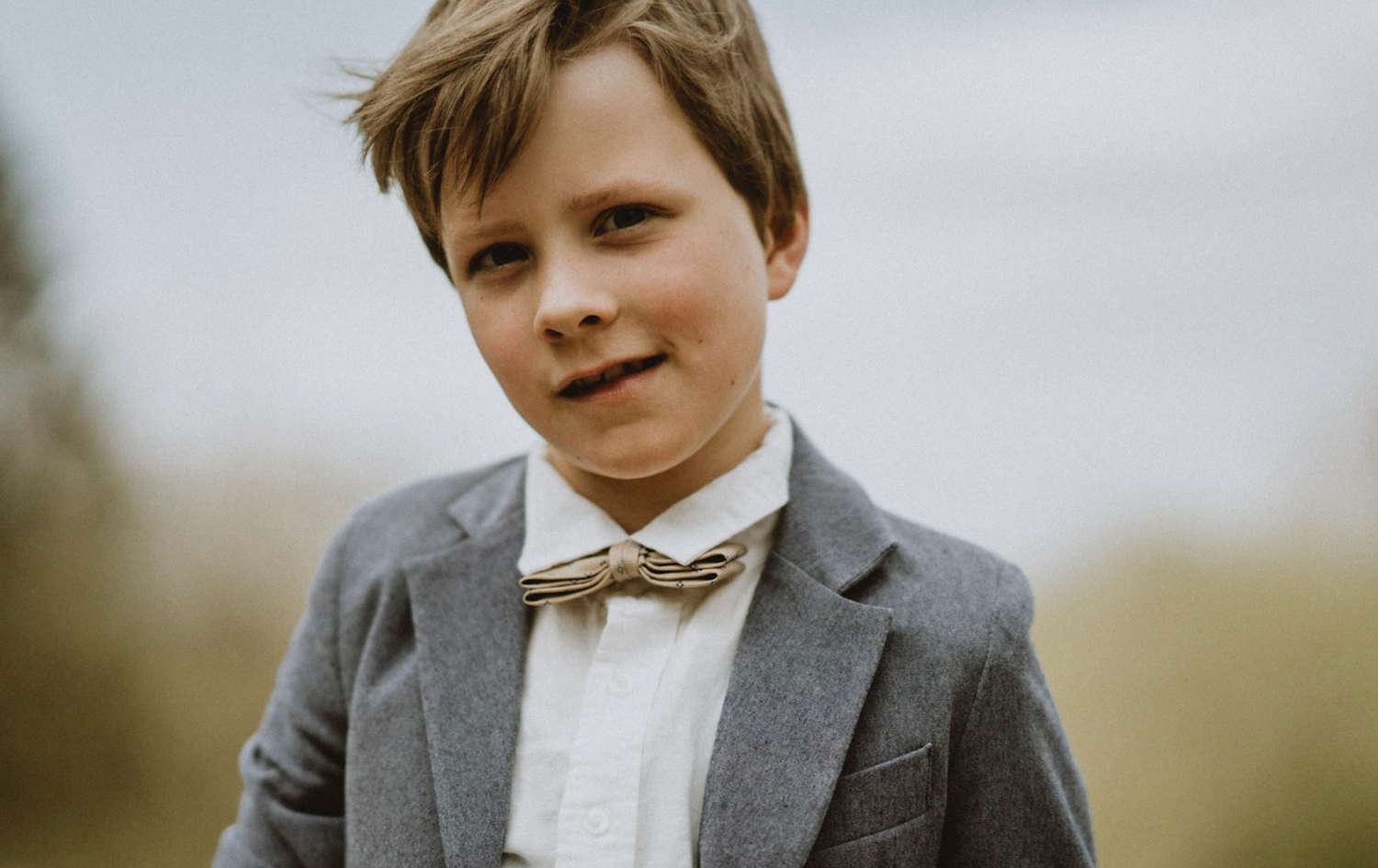 A portrait of a child wearing a grey suit jacket and bow tie against a blurred outdoor background.