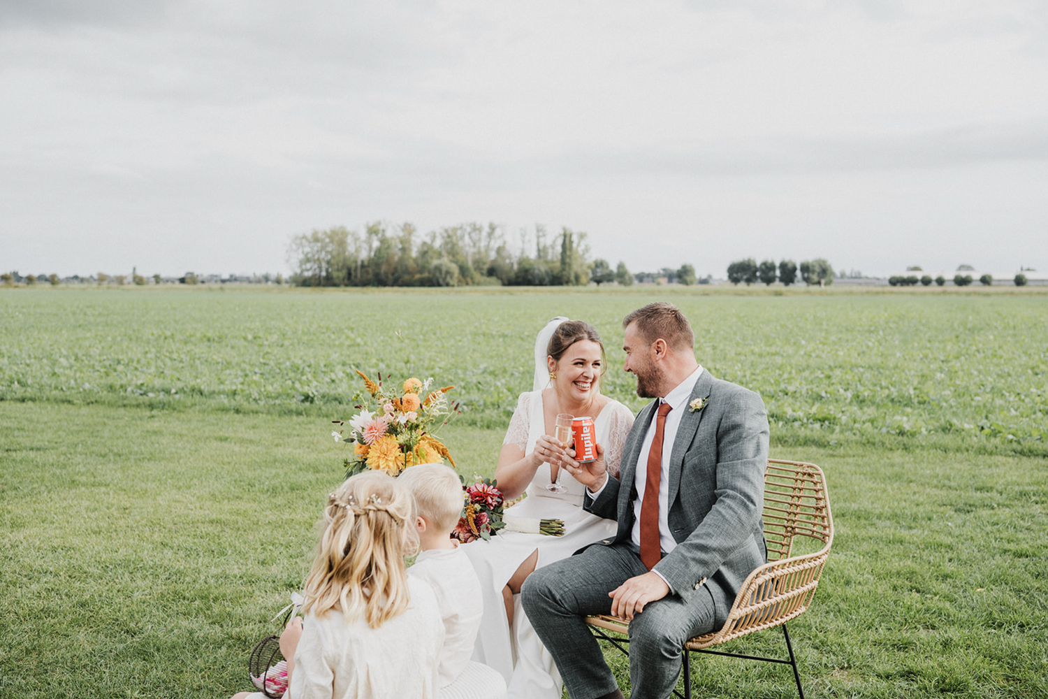 Bride and groom sitting on a rattan bench in a green field, sharing a romantic moment with colorful floral arrangements.