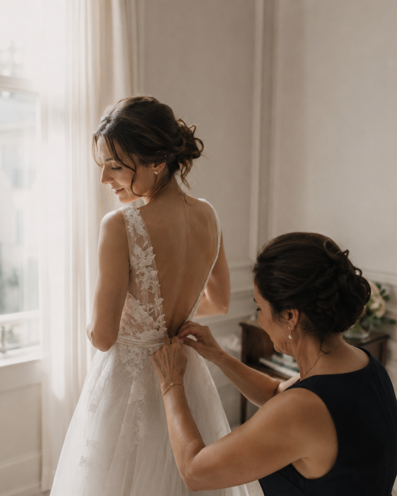 Bride in lace backless wedding gown being buttoned by bridesmaid near bright window.