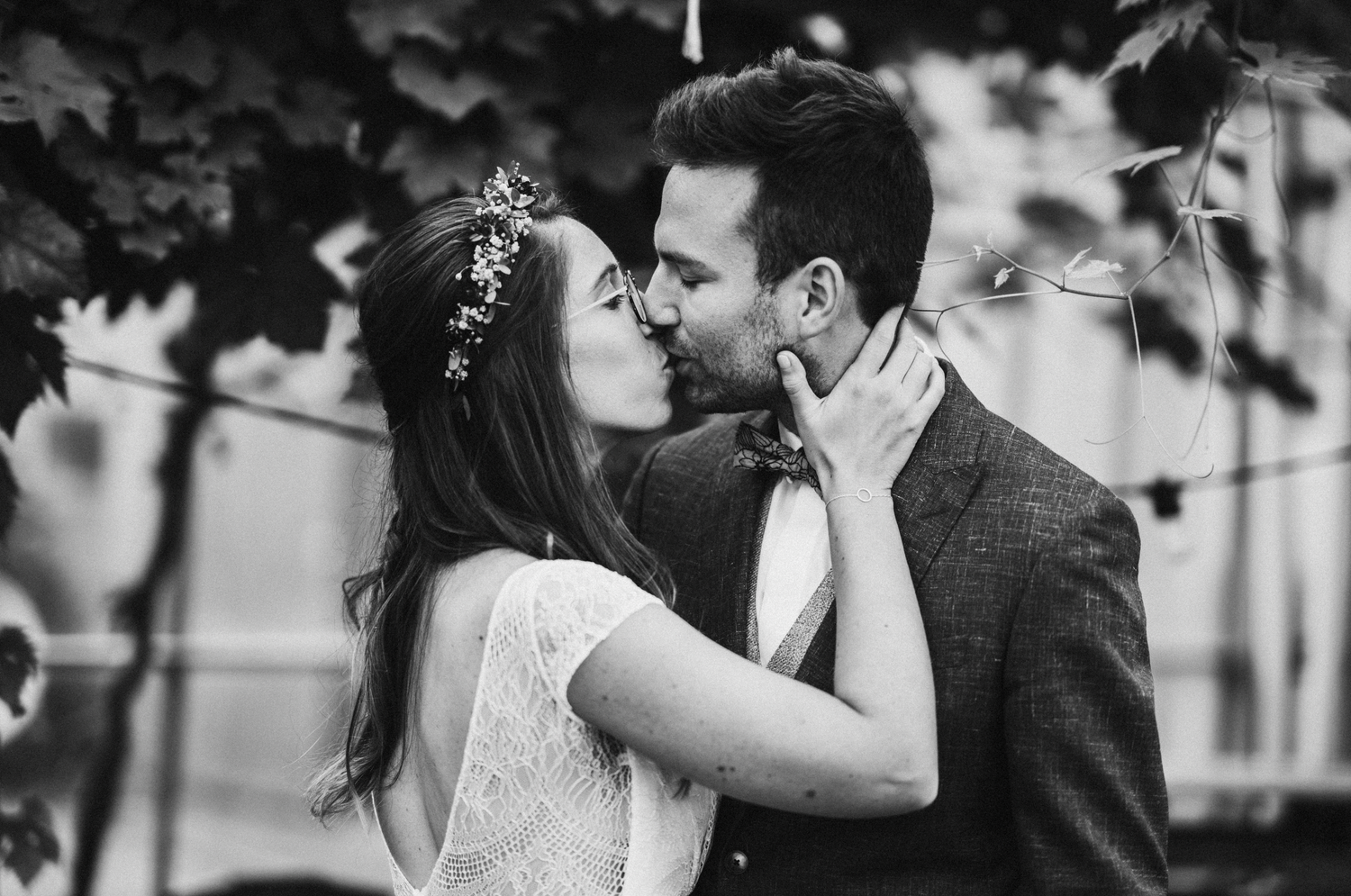 Black and white wedding photo of bride and groom sharing a romantic kiss outdoors among lush green leaves.