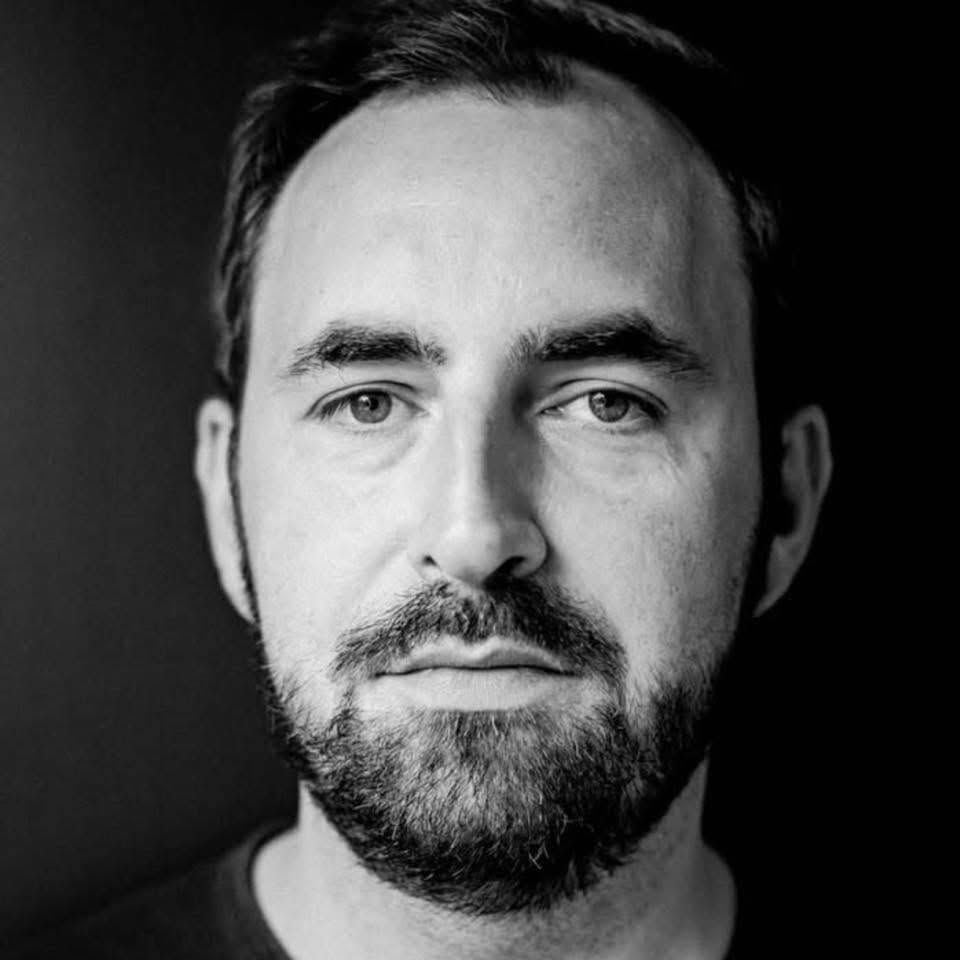 Black and white portrait of a bearded man with short hair, looking directly at the camera with a serious expression.