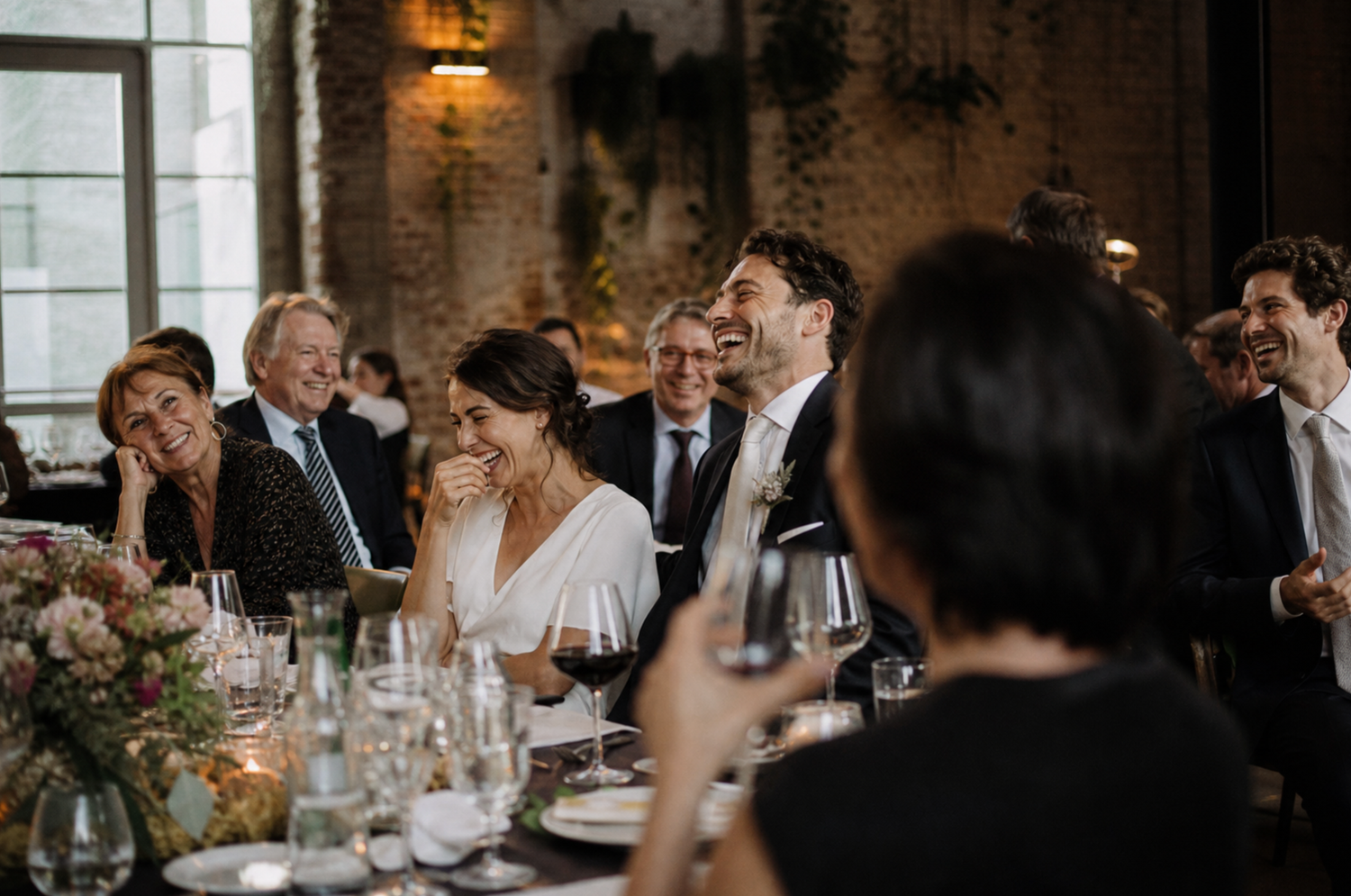 Bride and groom laughing joyfully at wedding reception dinner table surrounded by guests in elegant venue.