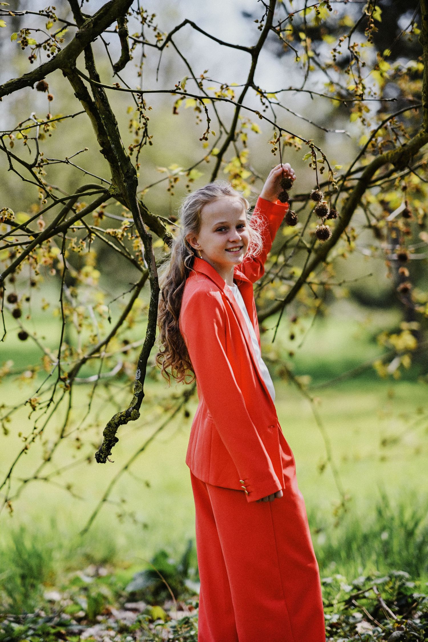 Young woman in red suit smiling beside a budding tree in a lush green garden.
