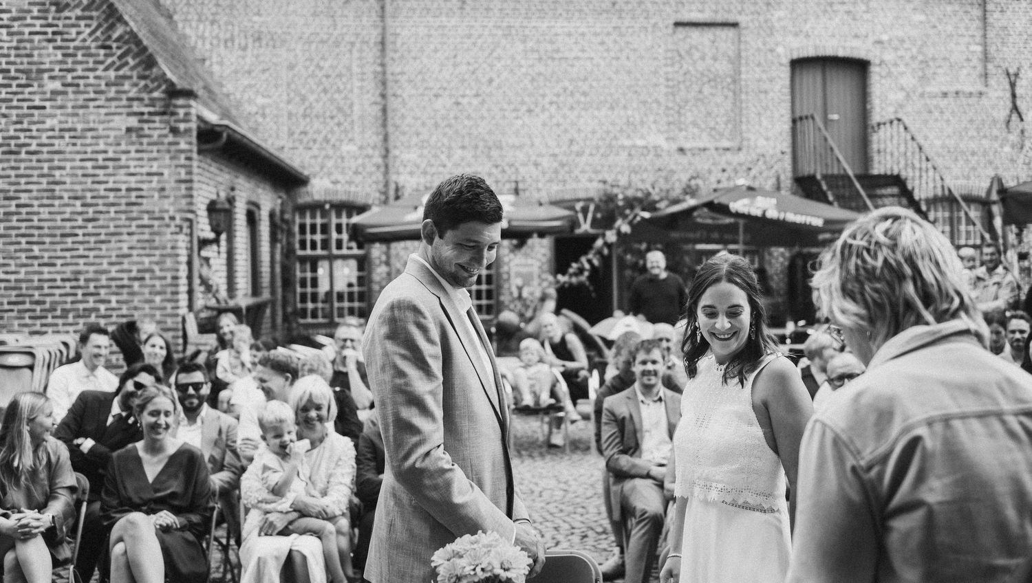 Black and white outdoor wedding ceremony with couple at altar, guests seated on chairs in rustic courtyard setting.