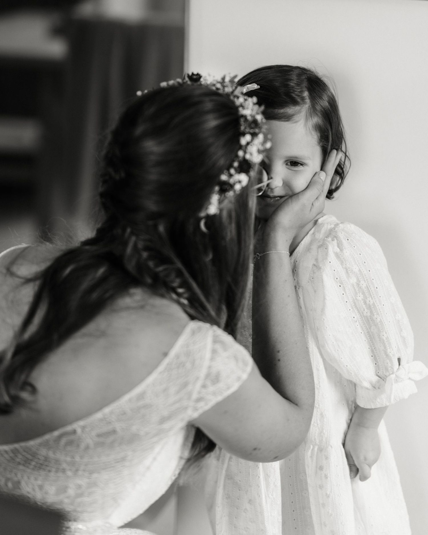 Black and white photo of a bride in lace dress tenderly cupping the face of a flower girl in a white dress.