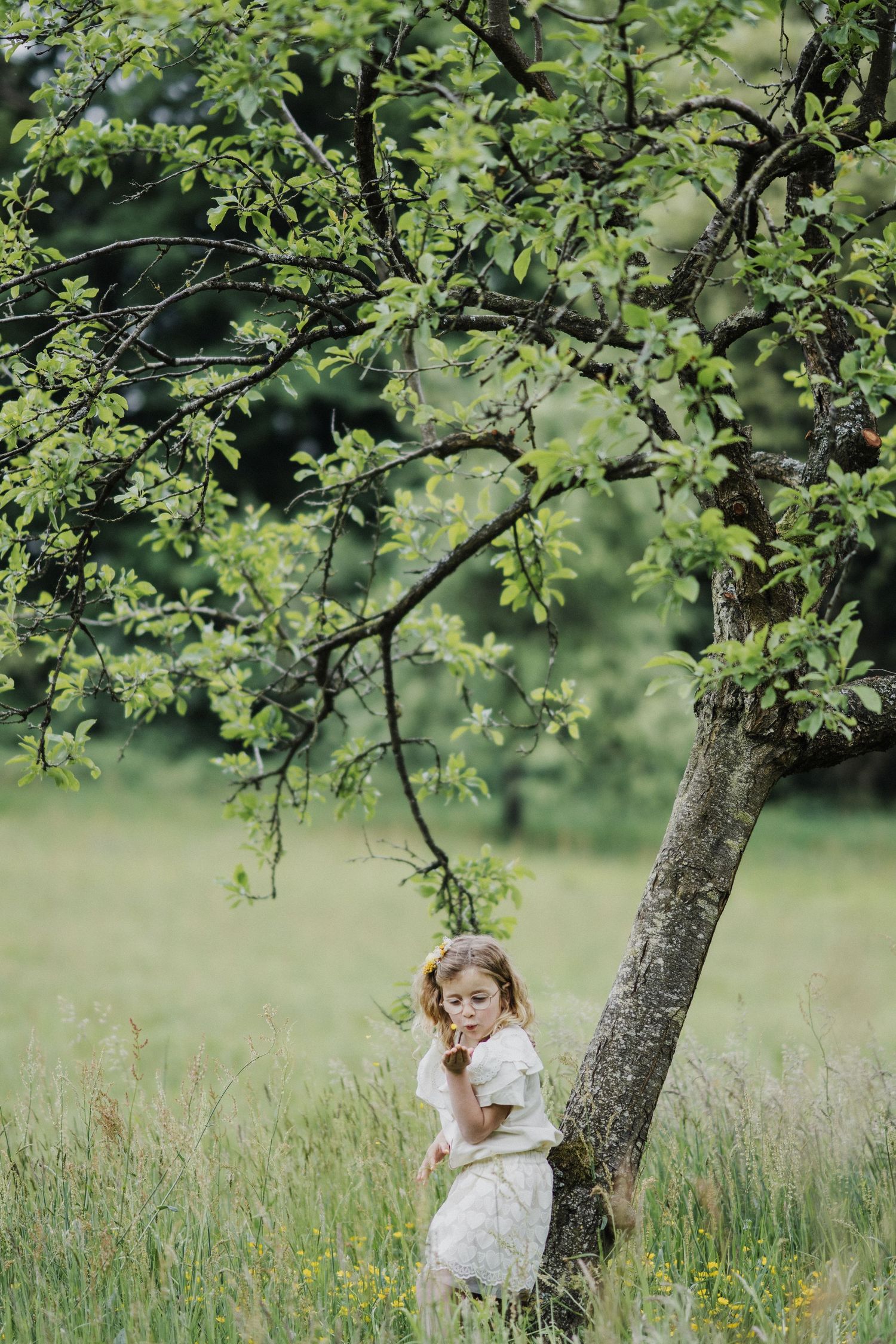 A young girl in a white dress stands beside a tree in a lush green meadow, holding a flower.