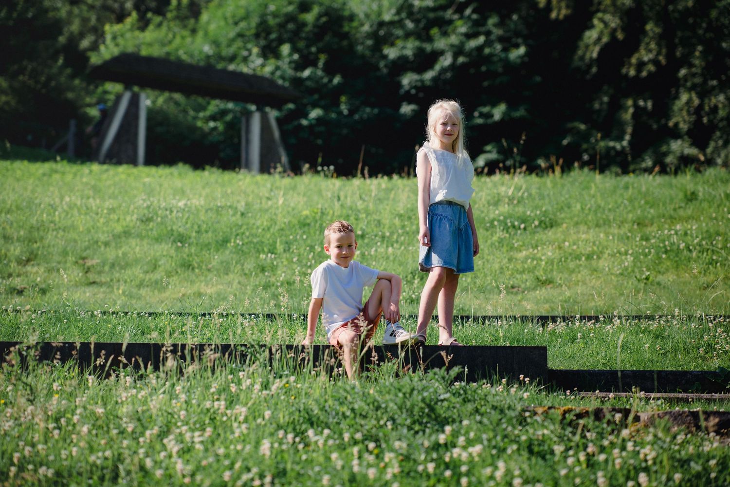Two blonde children playing outdoors on steps surrounded by lush green grass on a sunny day.