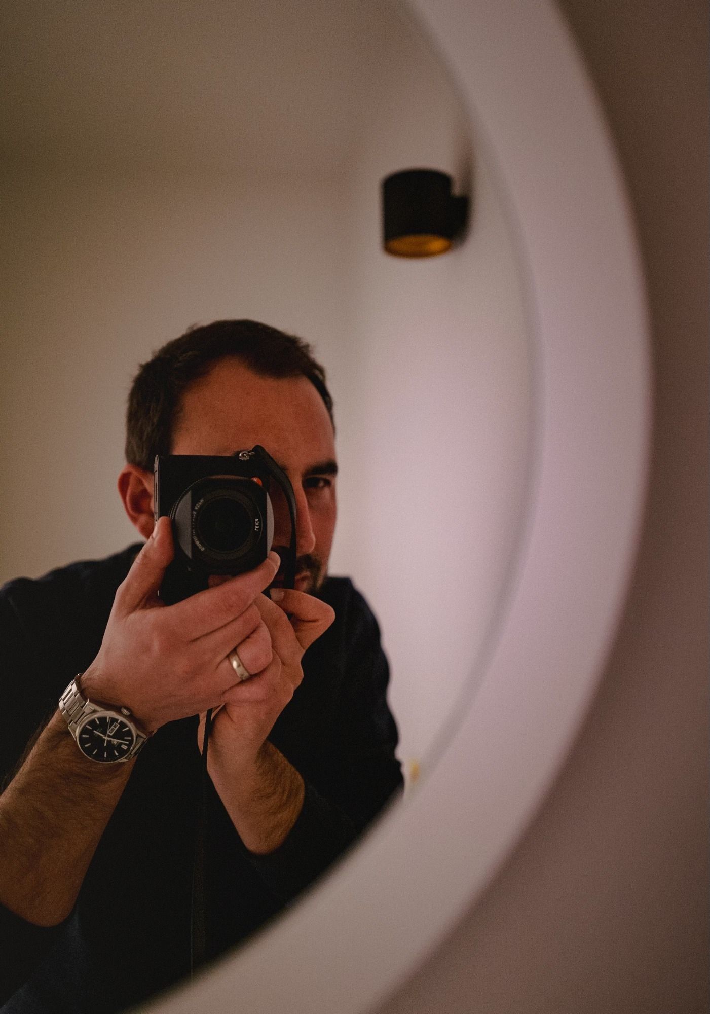 A photographer in dark clothing holds a camera to their eye in a dimly lit setting with circular light patterns.