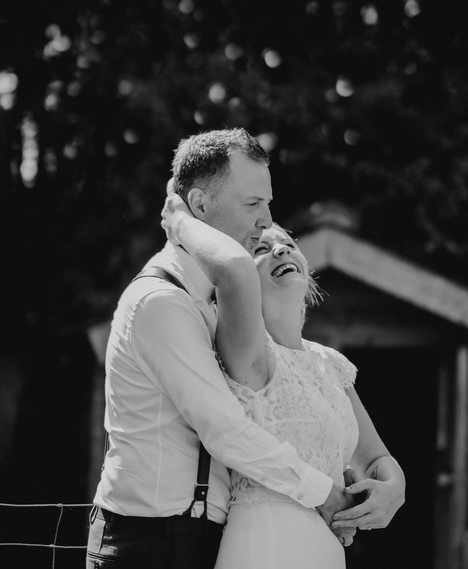 Black and white photo of a bride and groom embracing outdoors, laughing joyfully on their wedding day.