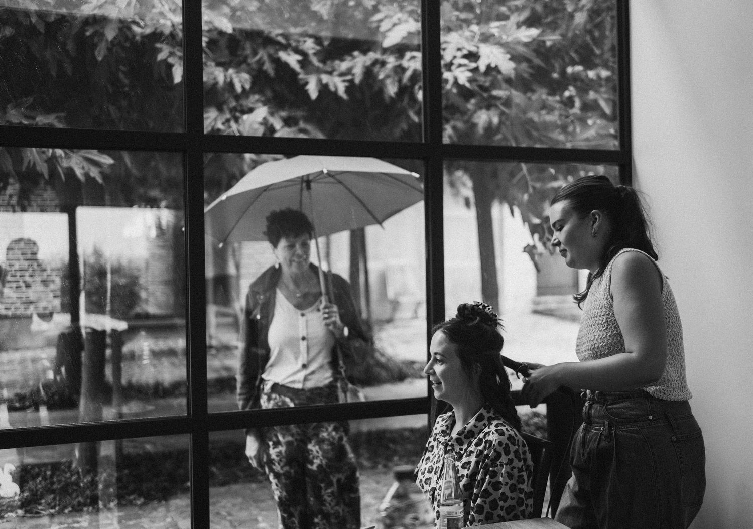 Black and white photo of women inside near large windows, one having hair styled while another holds umbrella outside.