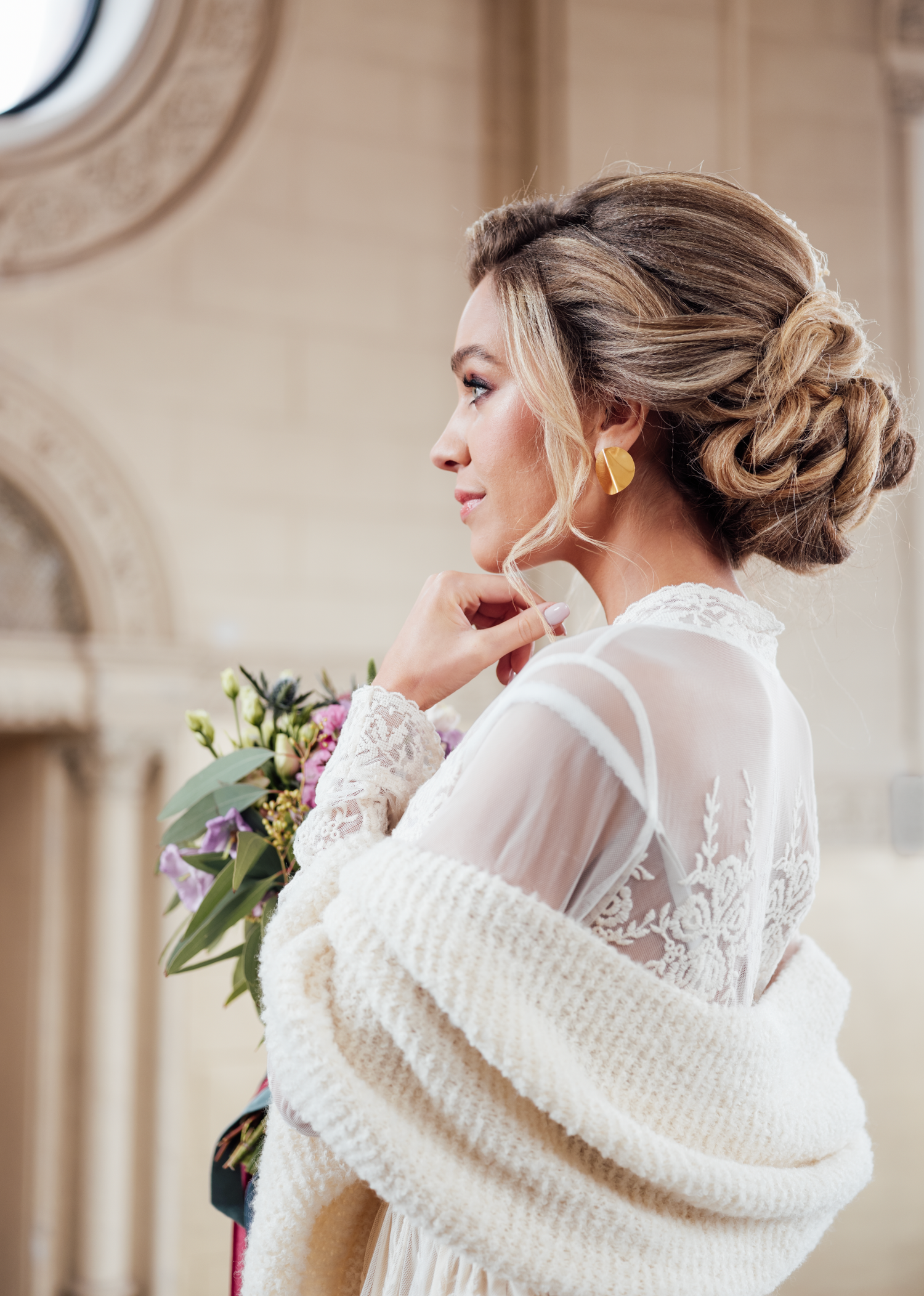 Elegant bride in lace gown with updo hairstyle holds floral bouquet, wearing gold earrings and white knit wrap.