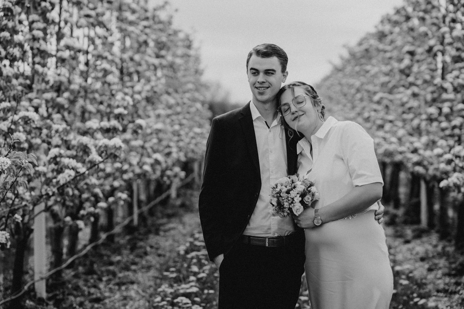 Black and white photo of a couple posing in a vineyard, woman holding a bouquet, both smiling warmly.