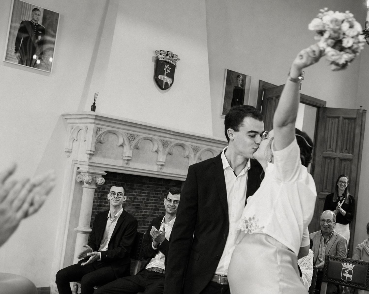 Black and white wedding photo of couple kissing while bride raises bouquet, guests seated in elegant room.