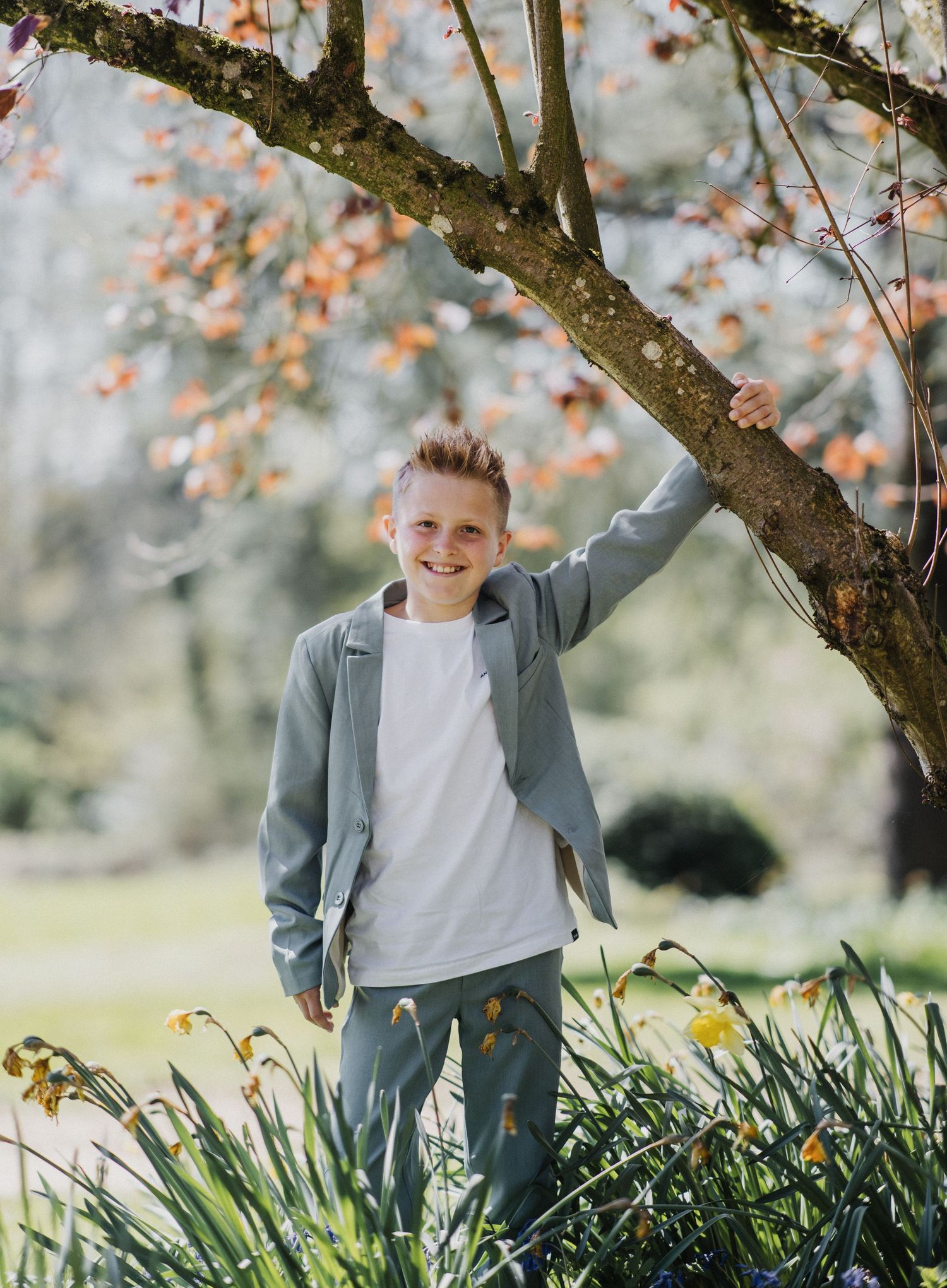 Smiling boy in sage green outfit standing under a flowering tree surrounded by daffodils in a lush spring garden.