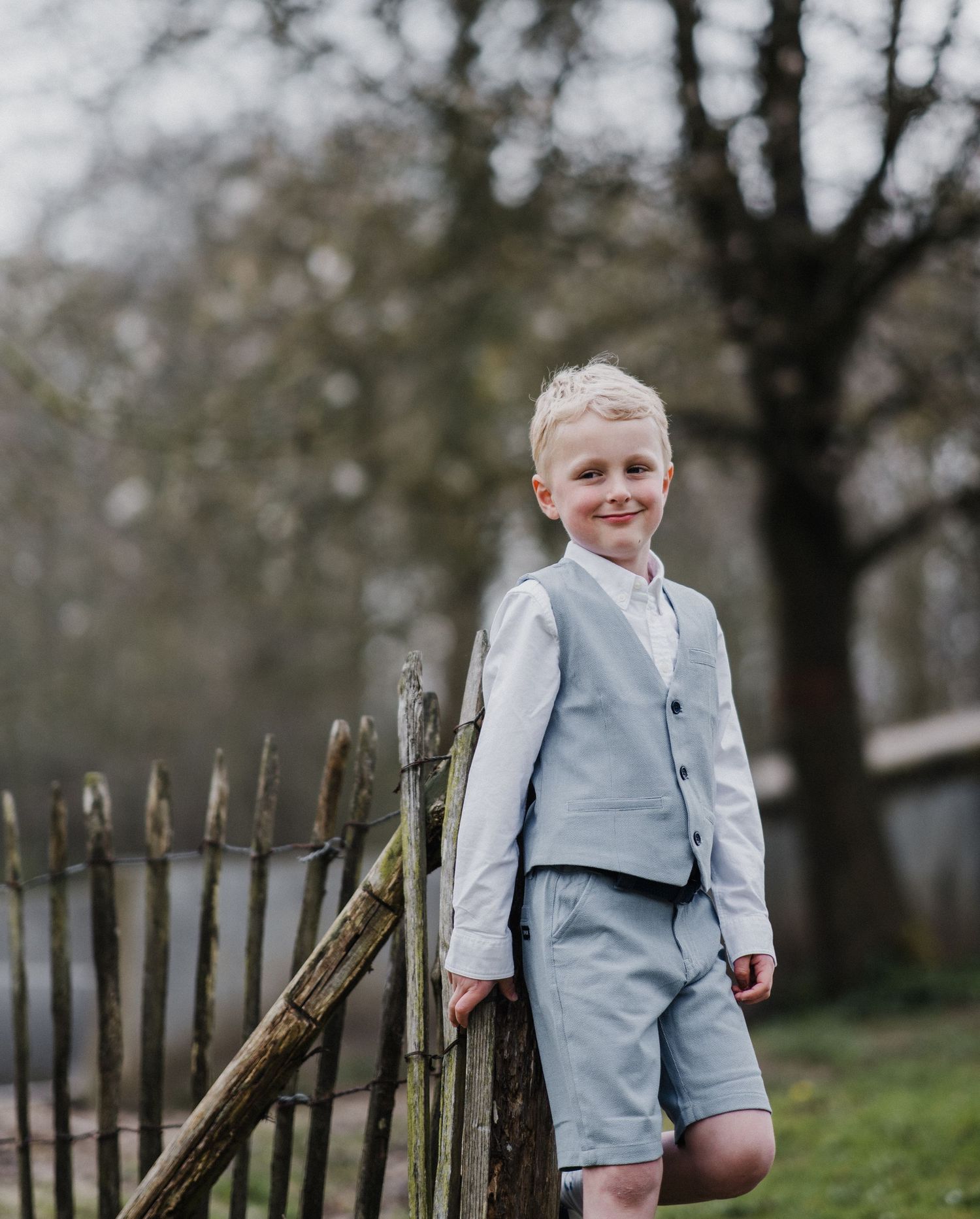 A young boy in a light blue suit and shorts smiles while leaning on a rustic wooden fence outdoors.