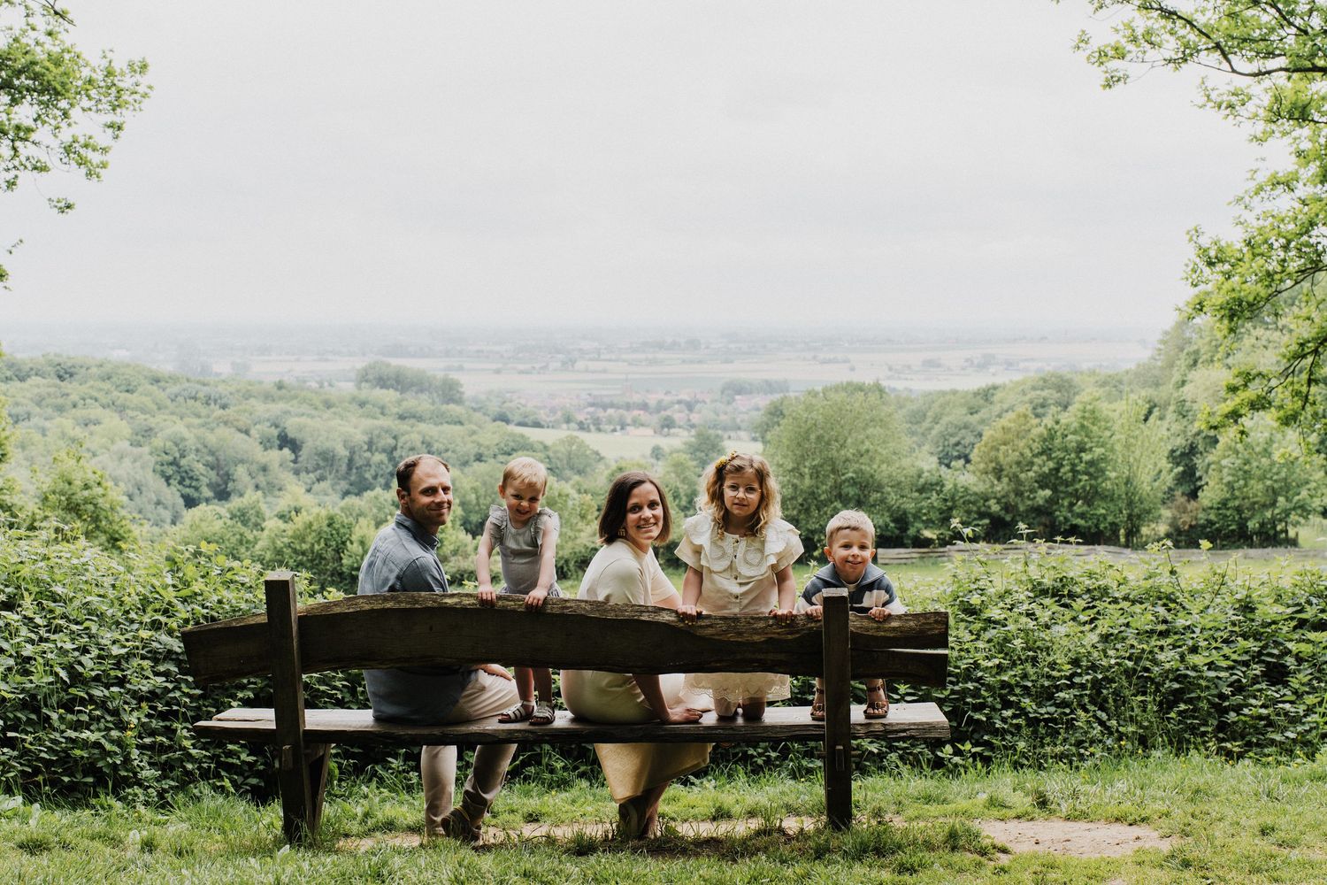 Family of five smiling together on a wooden bench surrounded by lush greenery with a scenic valley view behind them.