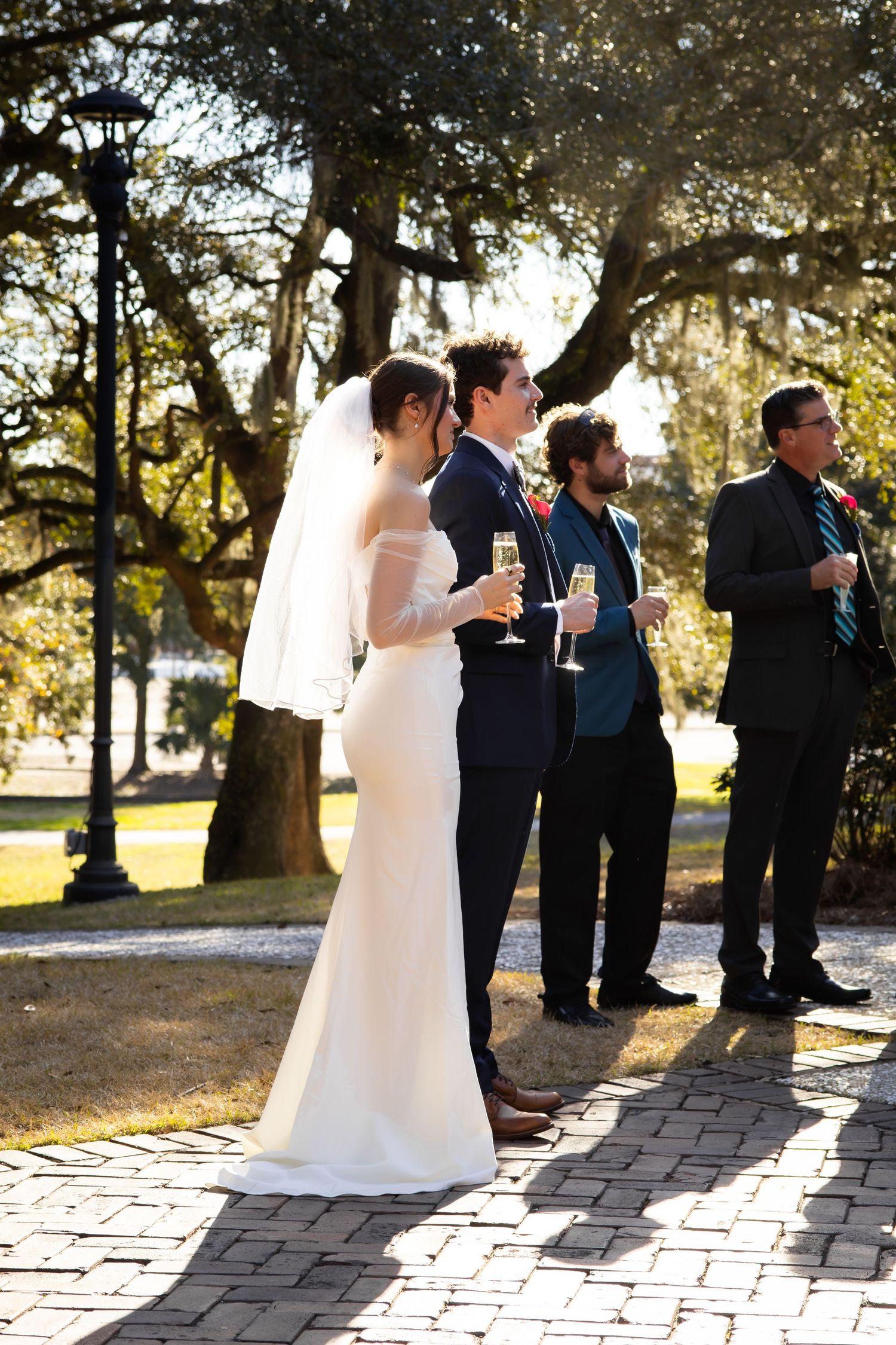 Wedding party lineup on brick pathway with trees casting shadows in afternoon light.