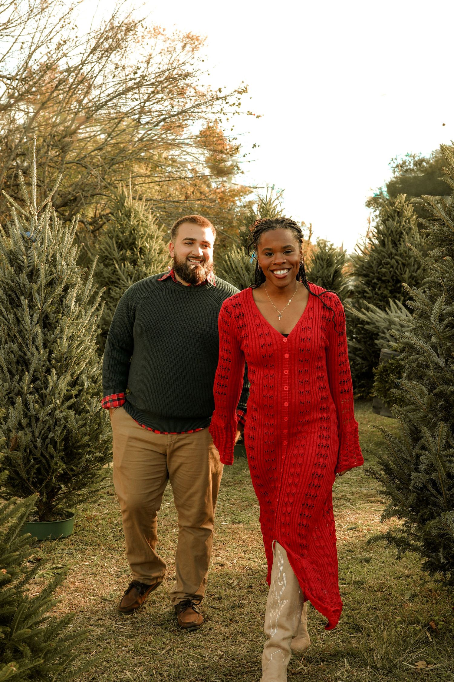 Couple walking through a Christmas tree farm at sunset, with one person wearing a festive red sweater dress.