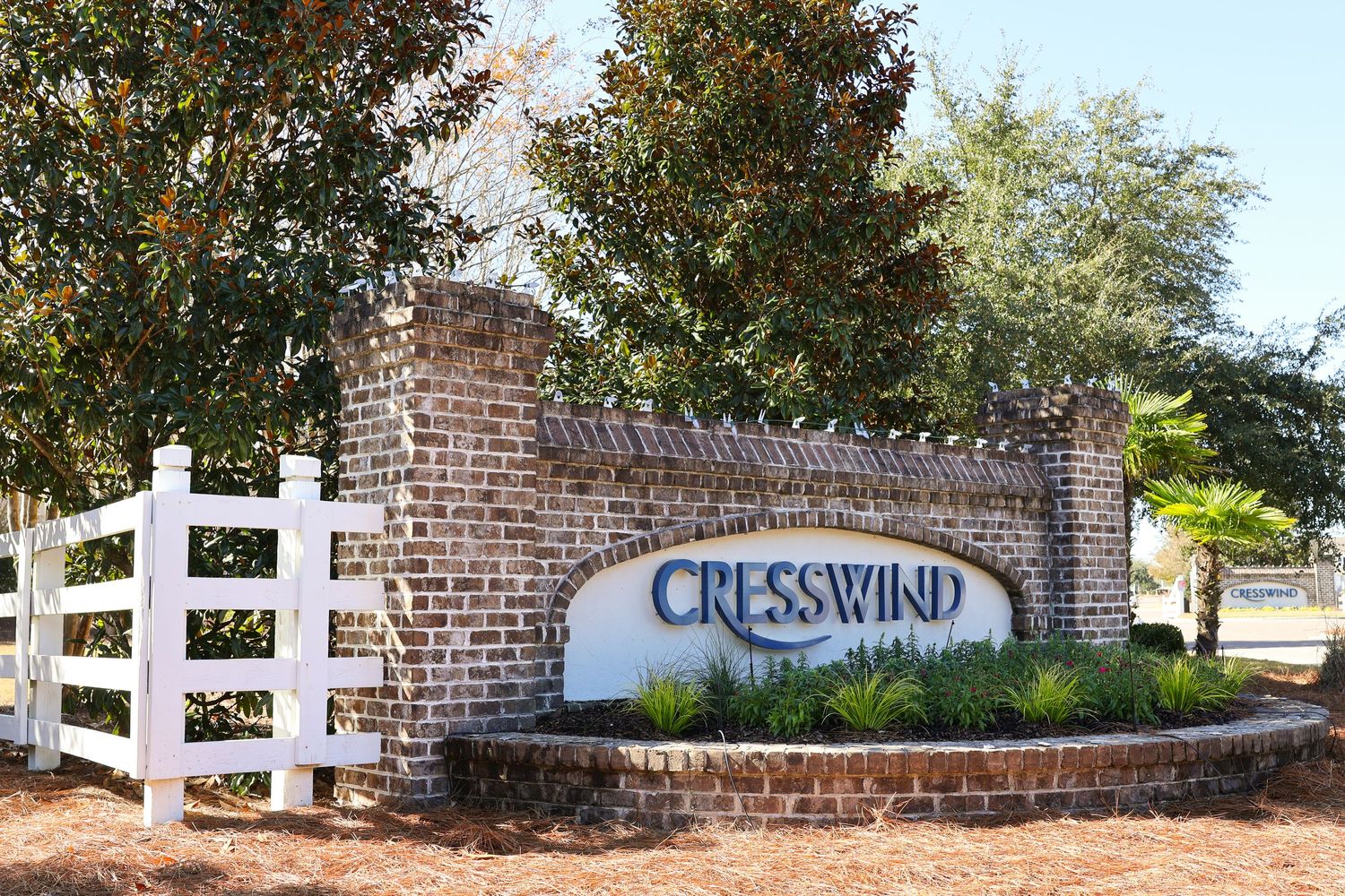 Brick entrance sign for Cresswind community with white fence and trees in background.