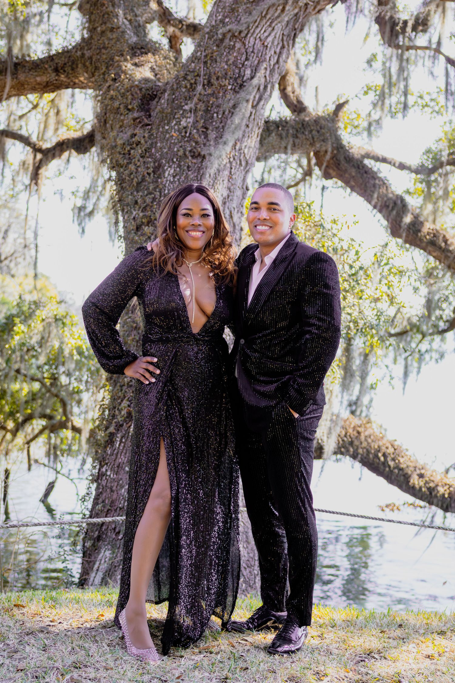 Couple poses playfully under moss-covered trees in formal black attire.