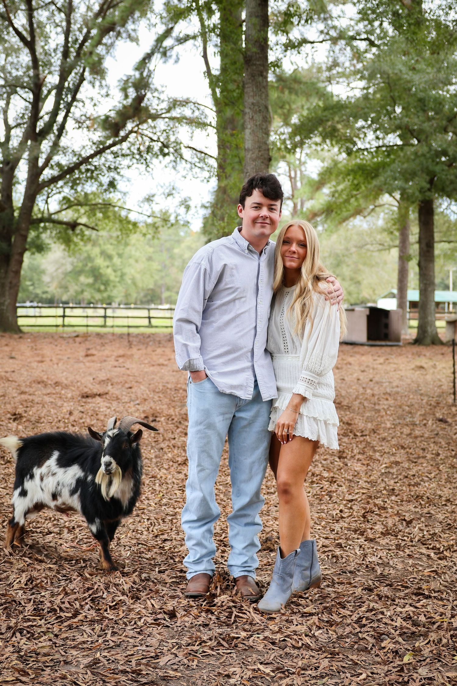 A couple poses with their black and white dog in a park setting with fallen leaves.