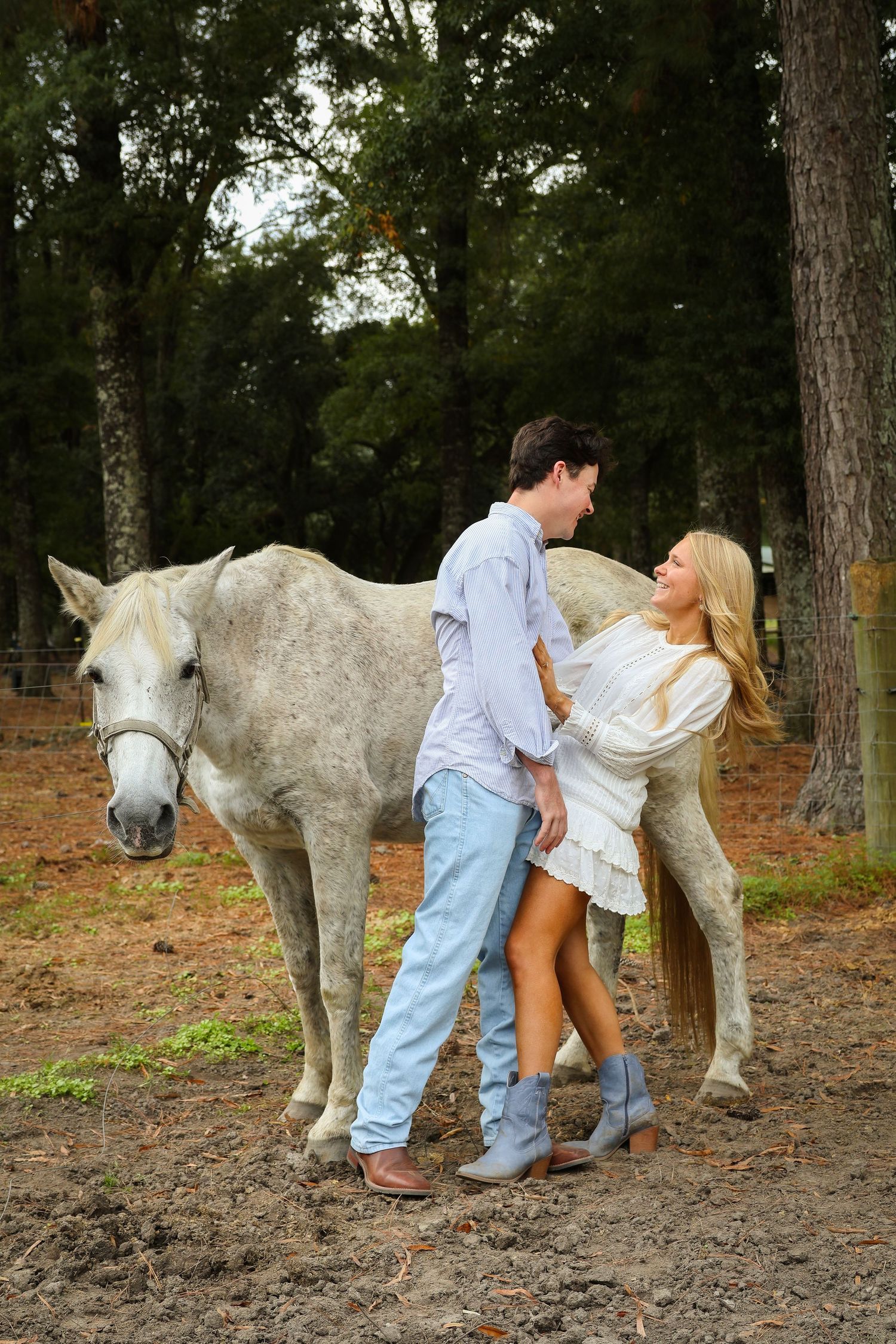 A couple poses playfully with a white horse in a wooded setting during a photo session.