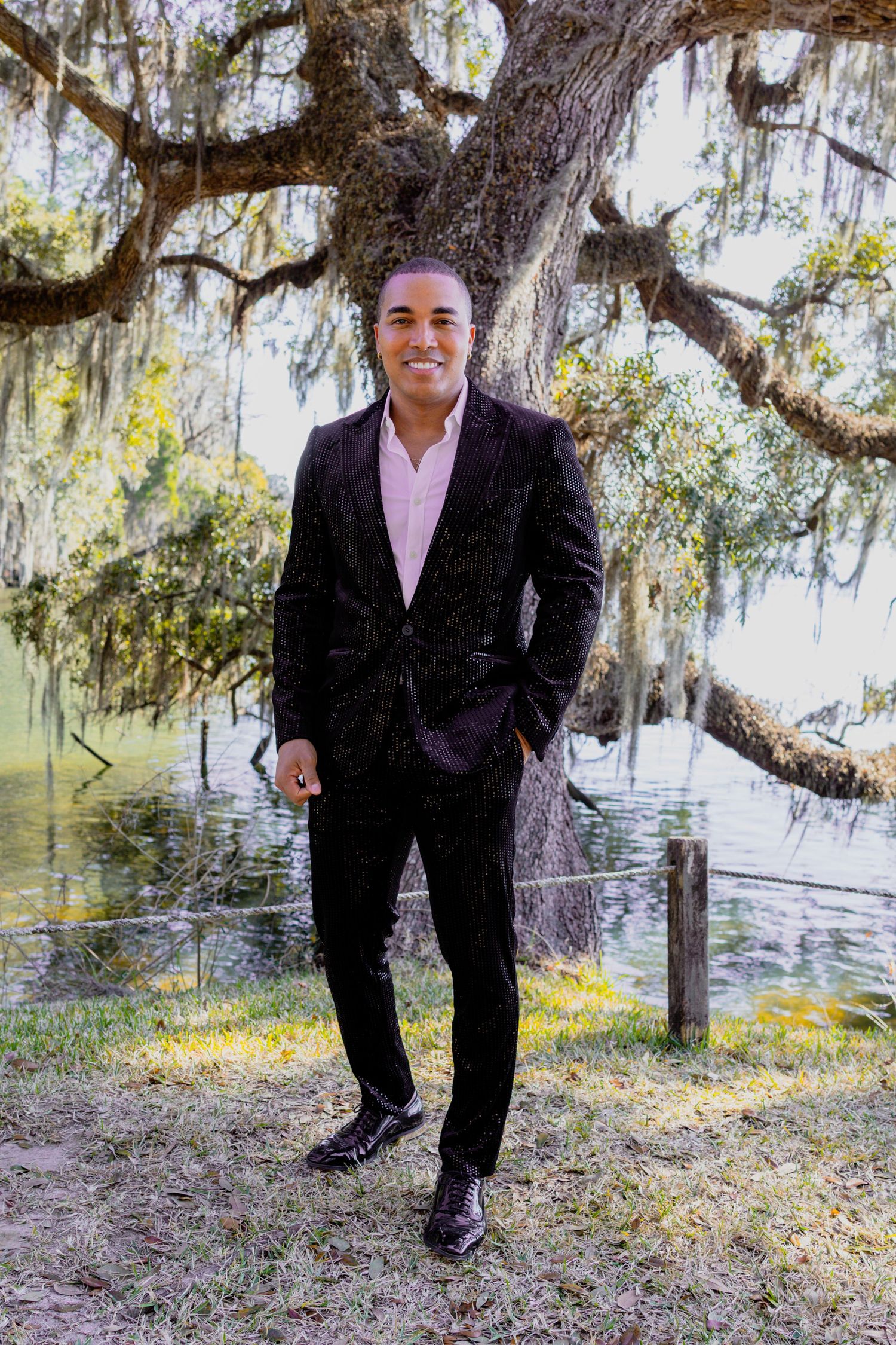 Person in business attire smiles while posing near water under Spanish moss trees.