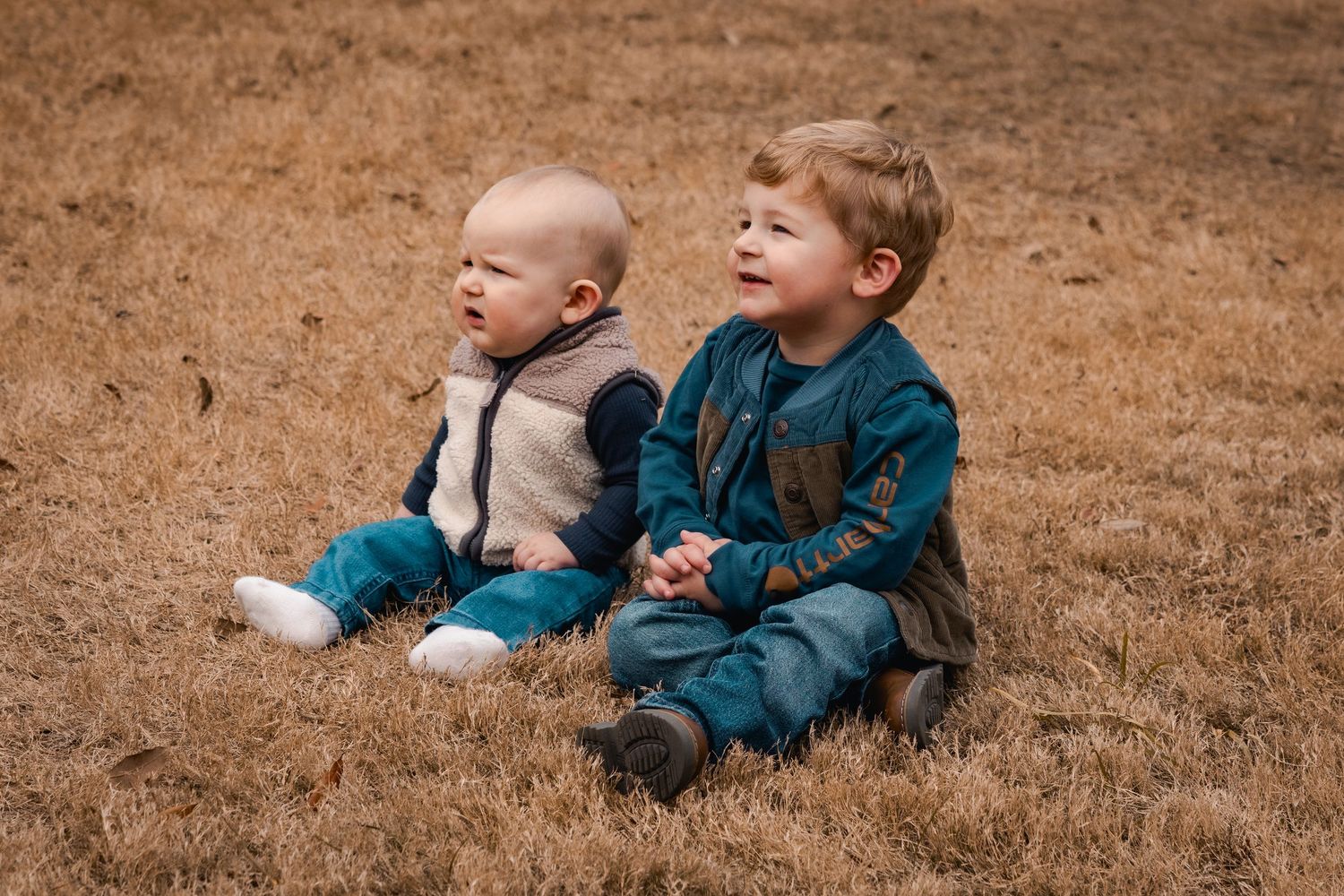 Two young children sit together on brown grass wearing coordinating blue and gray outfits.