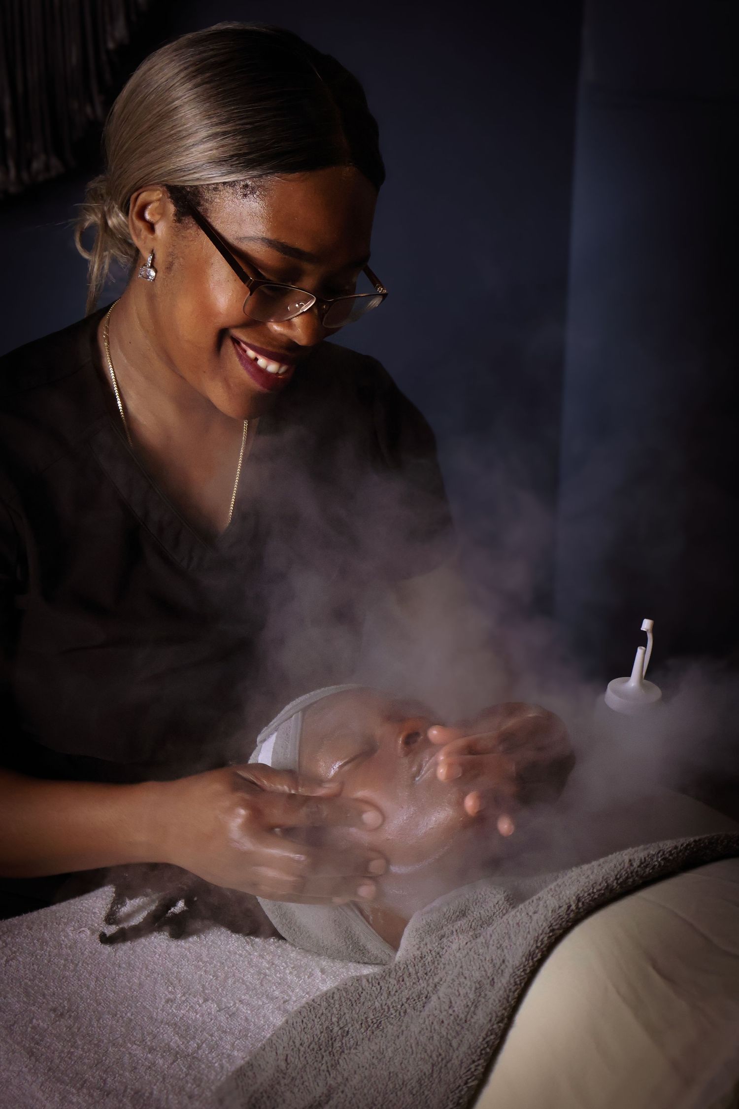 Esthetician performs facial treatment with steam in dramatic dark lighting.